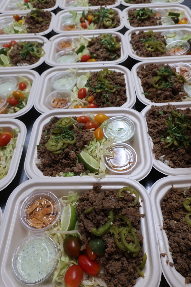 Multiple meal containers with ground meat, green peppers, cherry tomatoes, and small containers of sauce or dressing.