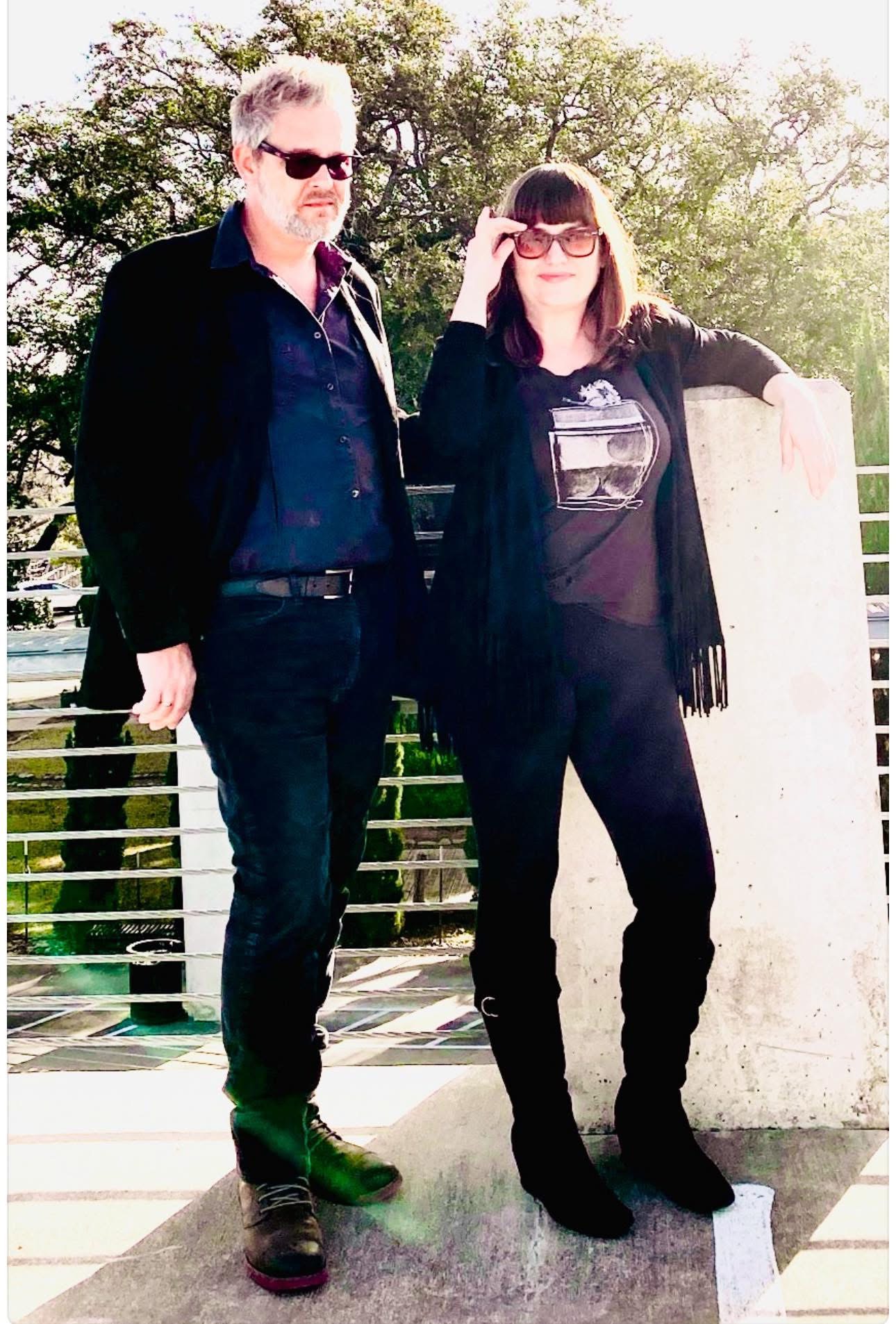 Two people standing outdoors, one man and one woman, both wearing sunglasses, with trees and a railing in the background.
