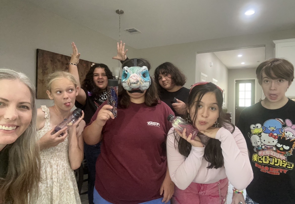 Group of seven people, some making peace signs, one wearing a mask, in a room with white walls and ceiling lights.