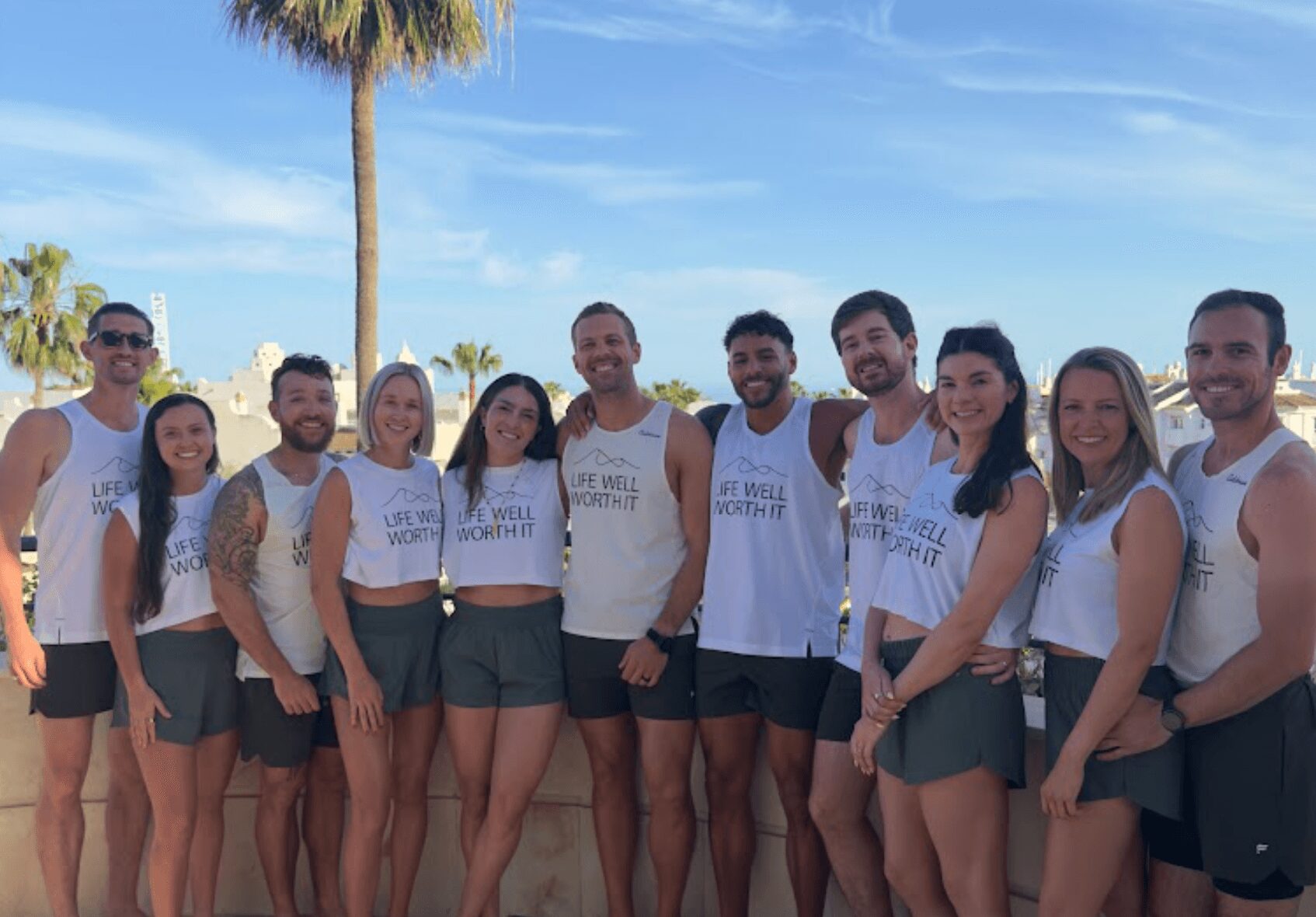 Group of ten people standing outdoors with palm trees and blue sky, smiling, wearing casual summer clothes.