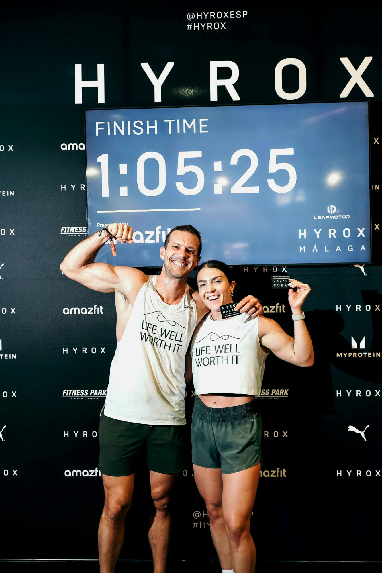 Two smiling runners holding a finish time sign of 1:05:25, standing in front of a black backdrop with white text.