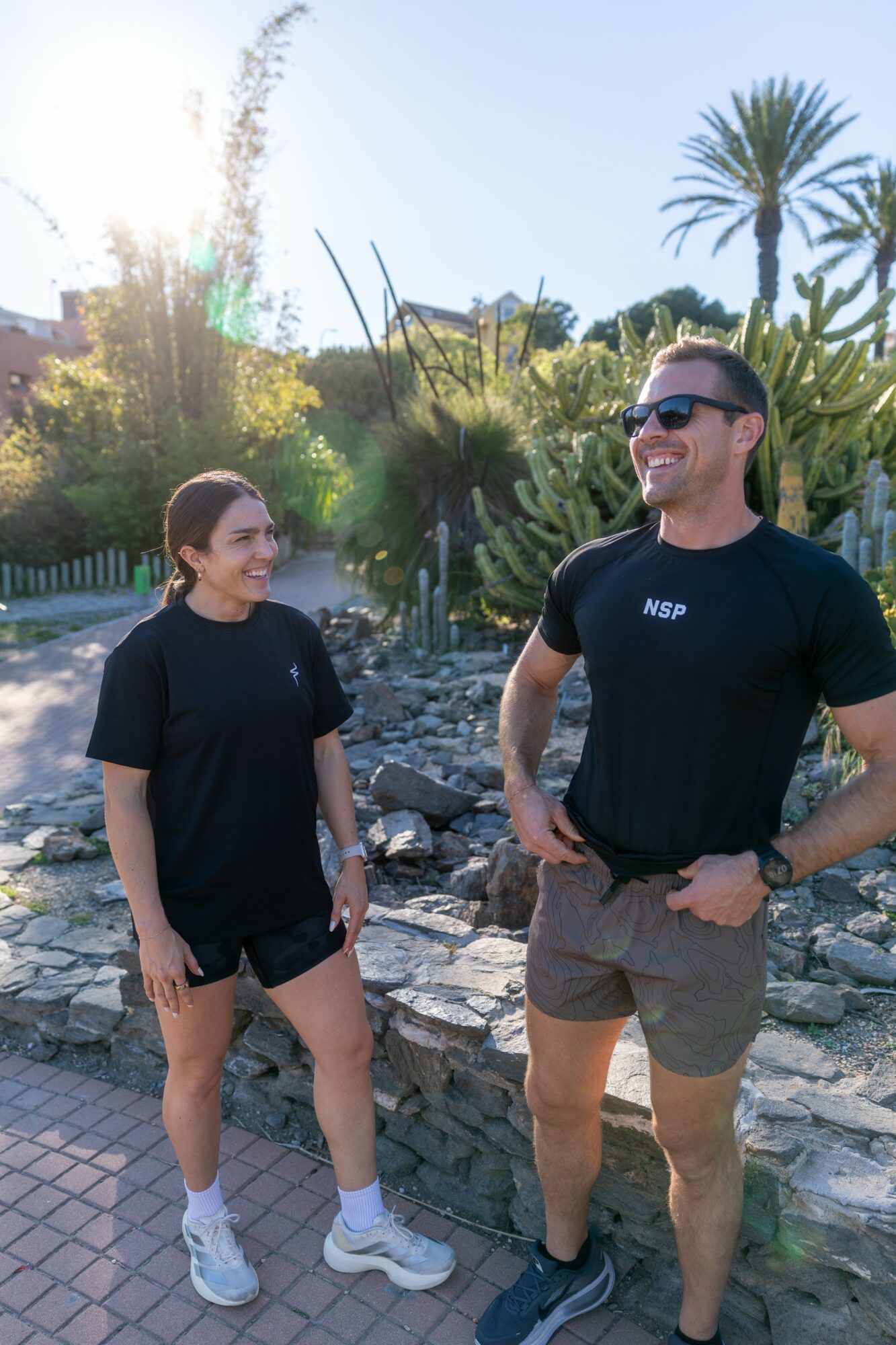 Two people, a woman and a man, stand outdoors near a rocky area with plants and trees, smiling and talking.