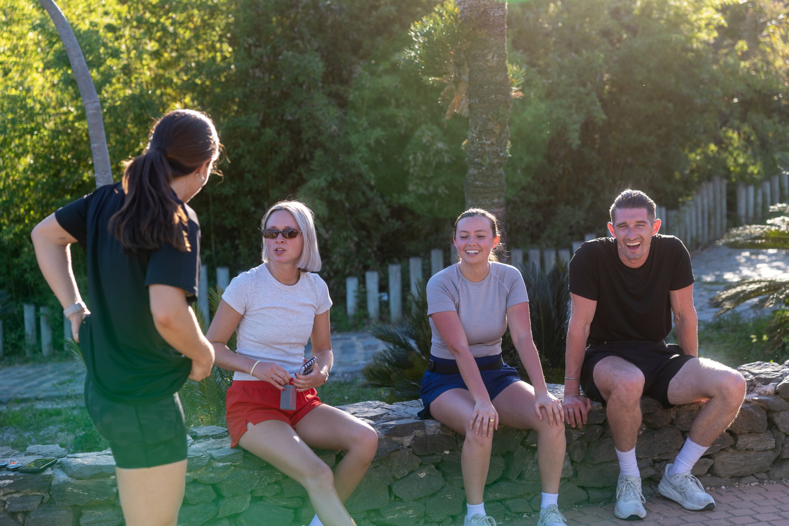 Four people sitting outdoors on a stone wall, smiling and talking, with trees and sunlight in the background.