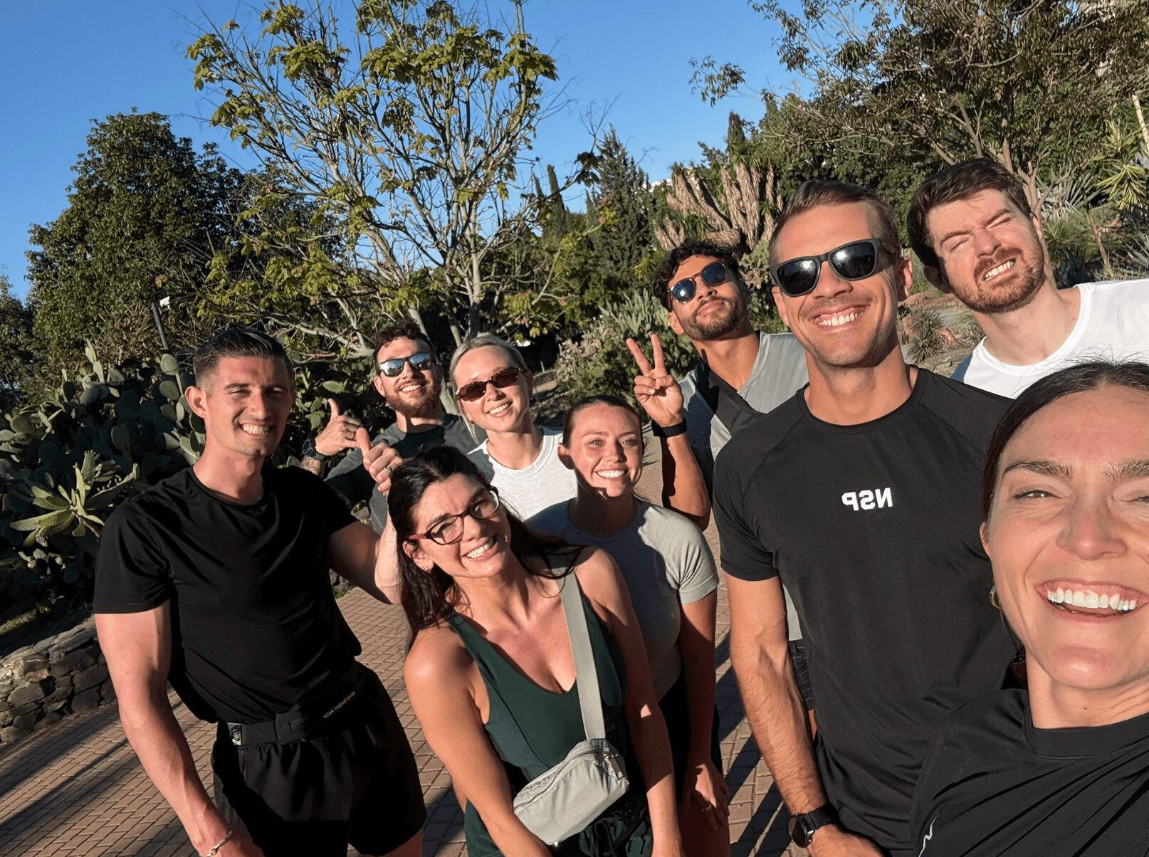 Group of nine people smiling outdoors with trees and blue sky in background.