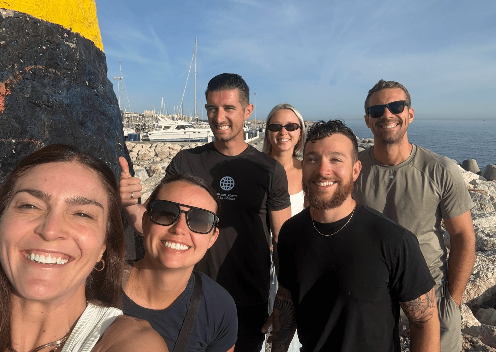 Group of six people smiling outdoors near a rocky shoreline with boats in the background.