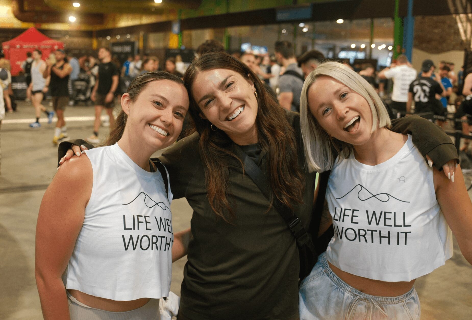 Three smiling women standing close together at an indoor event, with people and booths in the background.