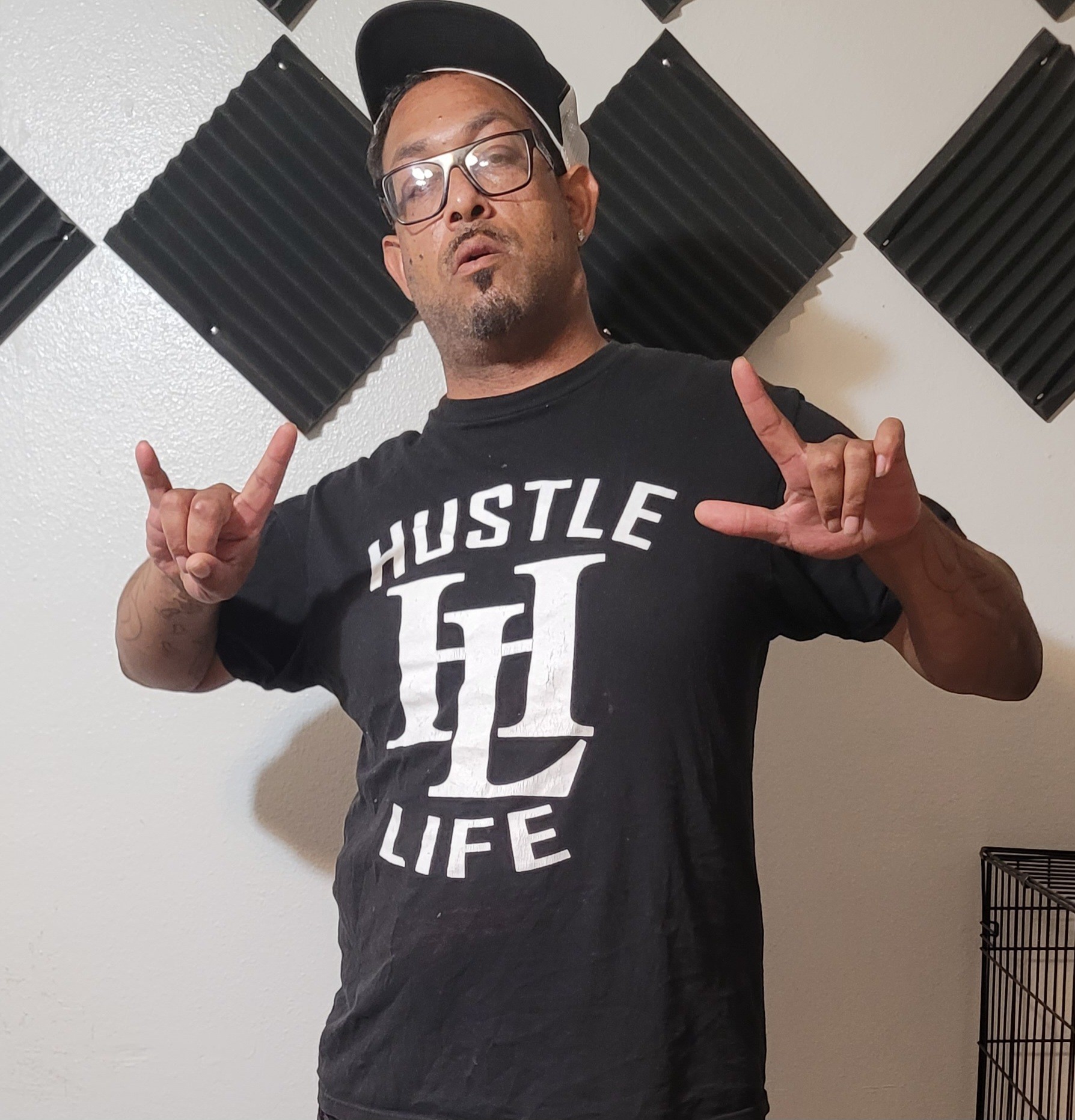 Man wearing glasses and a cap making hand signs, standing in front of soundproofing panels, smiling slightly.