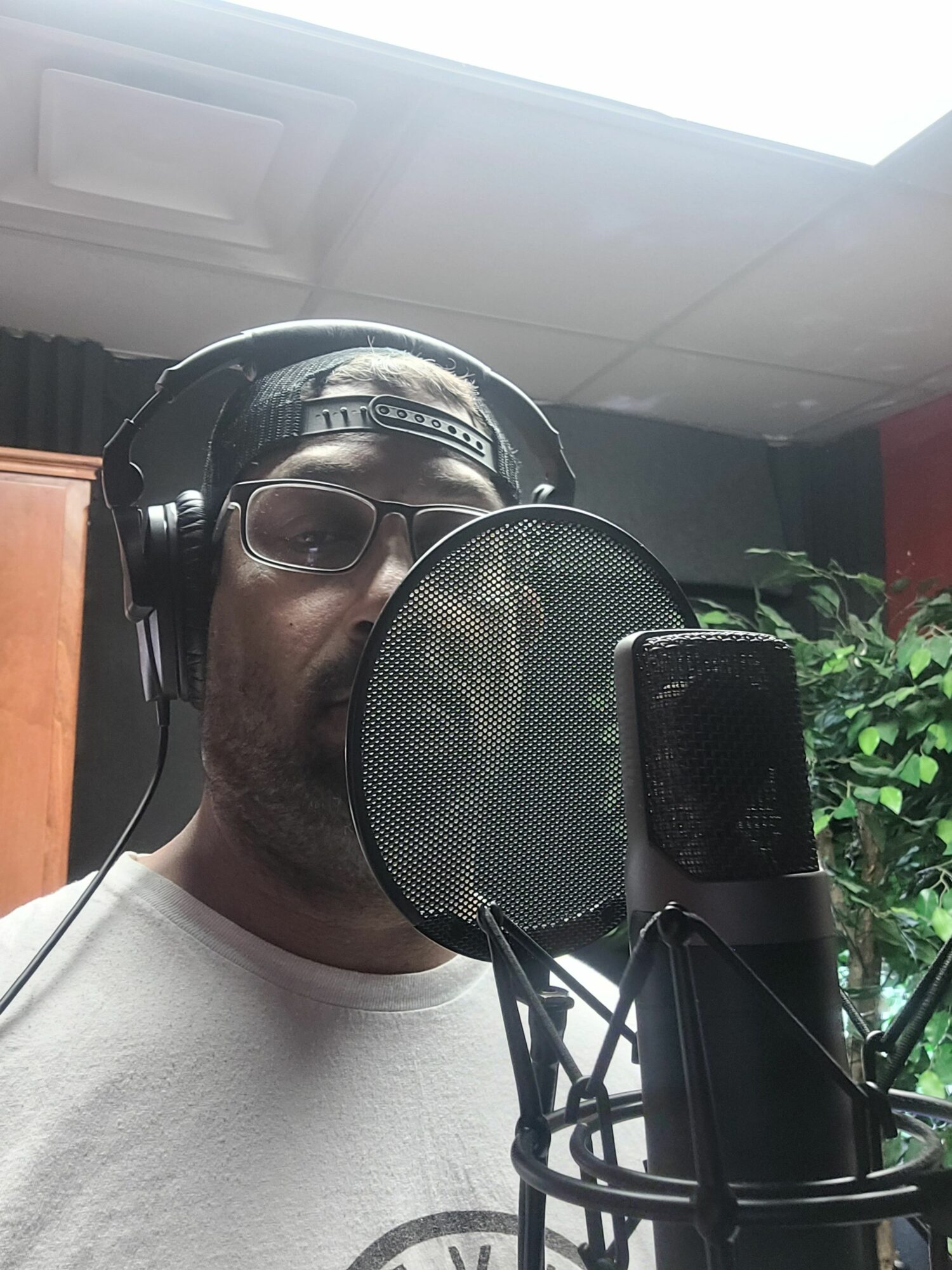 Person wearing headphones and glasses speaking into a microphone in a recording studio, with green plants in the background.
