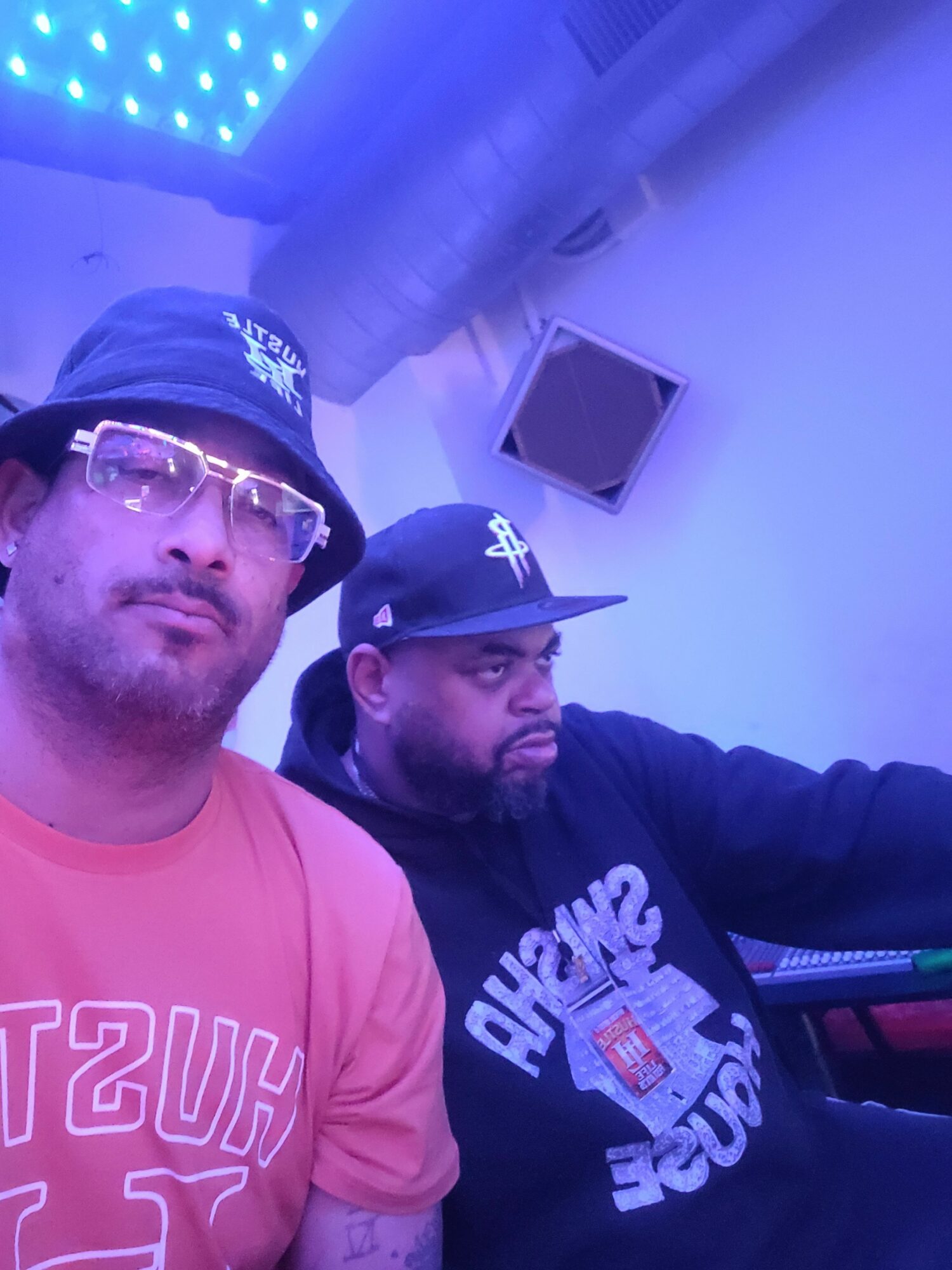 Two men pose indoors, one wearing a baseball cap and glasses, the other wearing a cap and hoodie, with blue lighting.