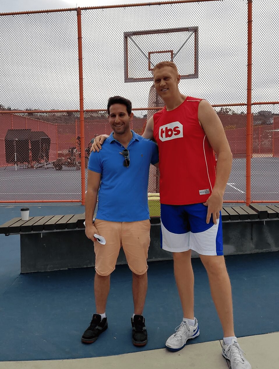 Two men stand on a basketball court, one in a blue shirt and beige shorts, the other in a red basketball jersey and blue shorts, smiling.