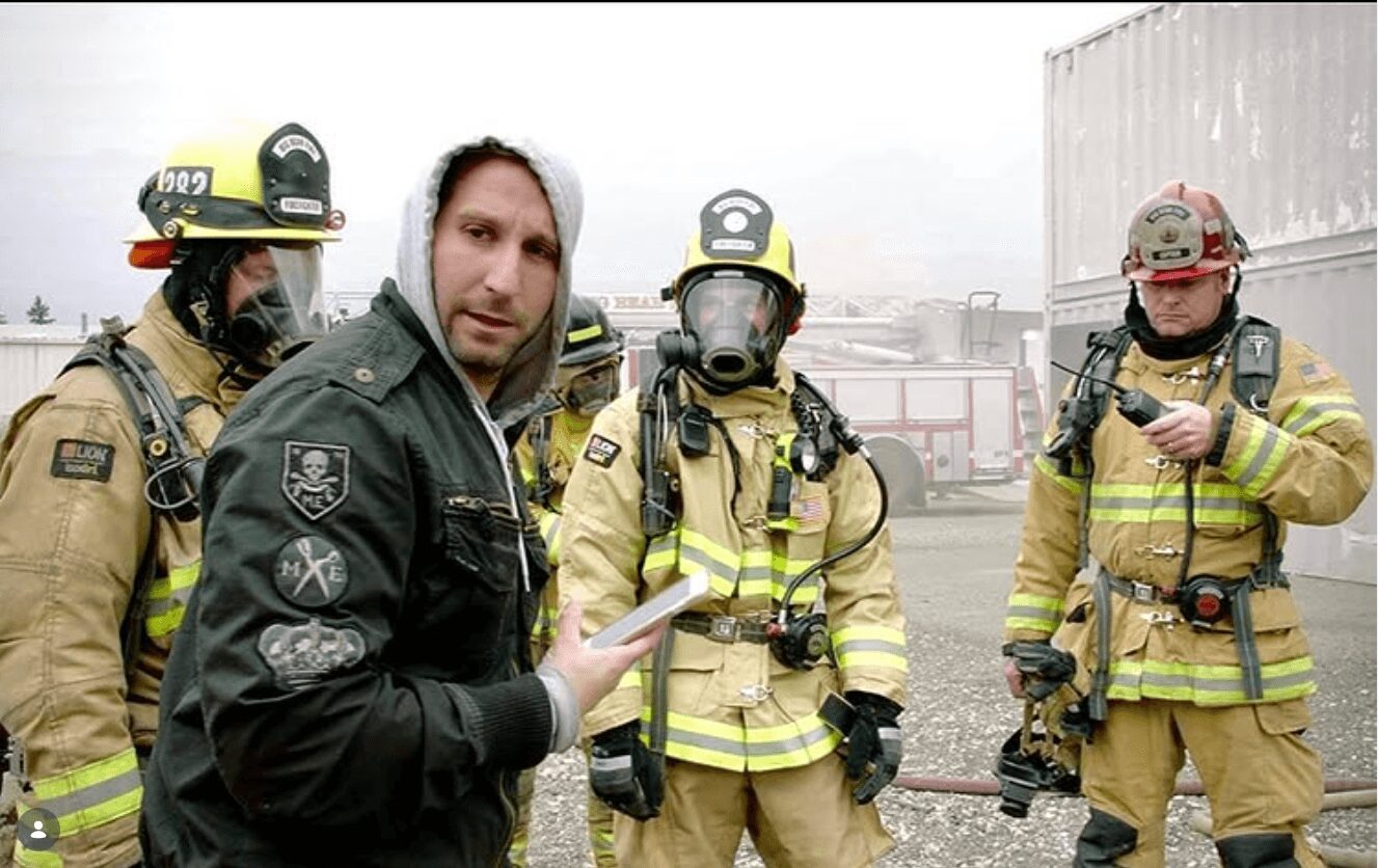 Man speaking to firefighters in protective gear outdoors, smoke and industrial buildings in background.