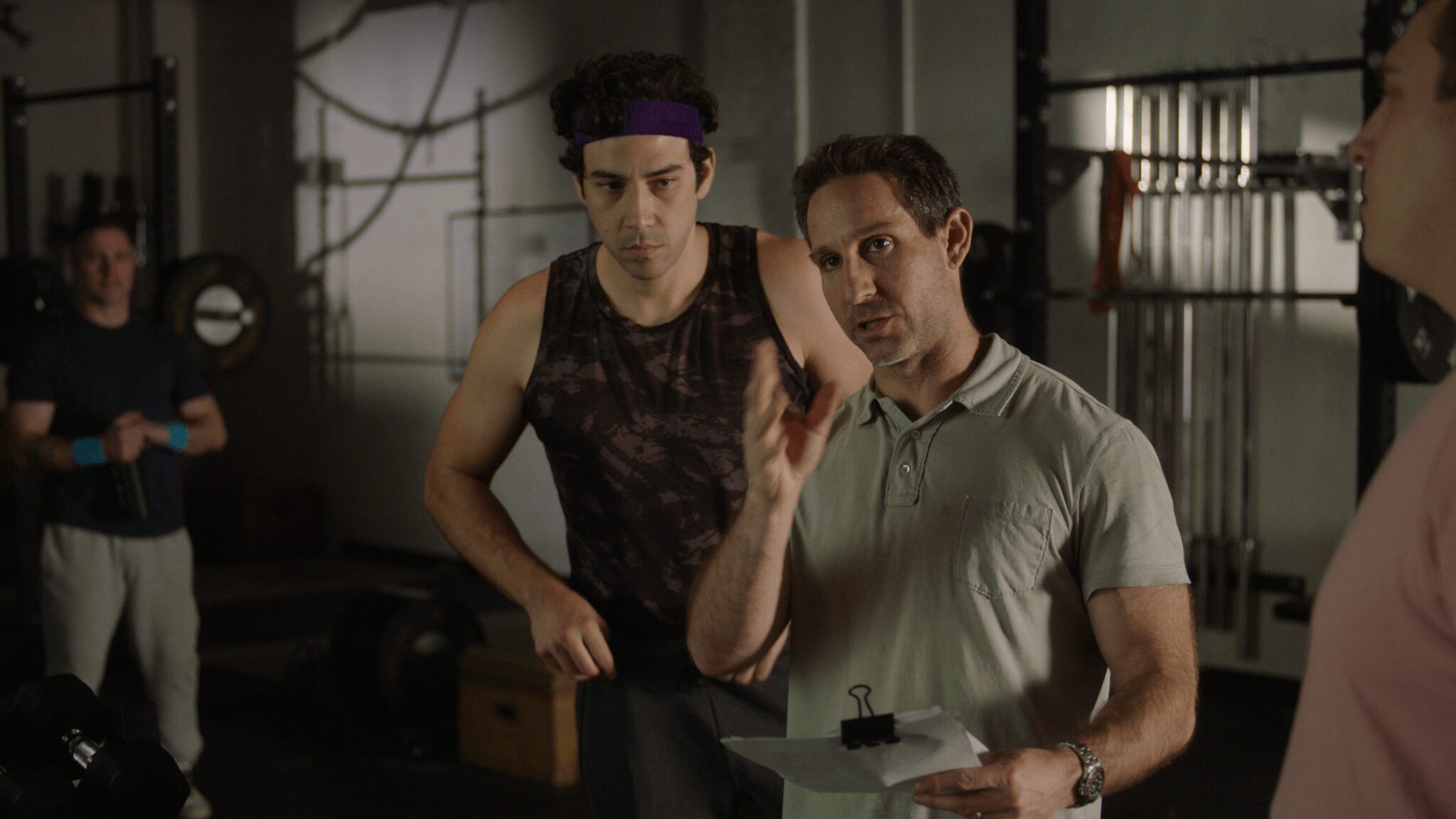 Two men in a room, one speaking and gesturing, others in the background, industrial setting, dim lighting.