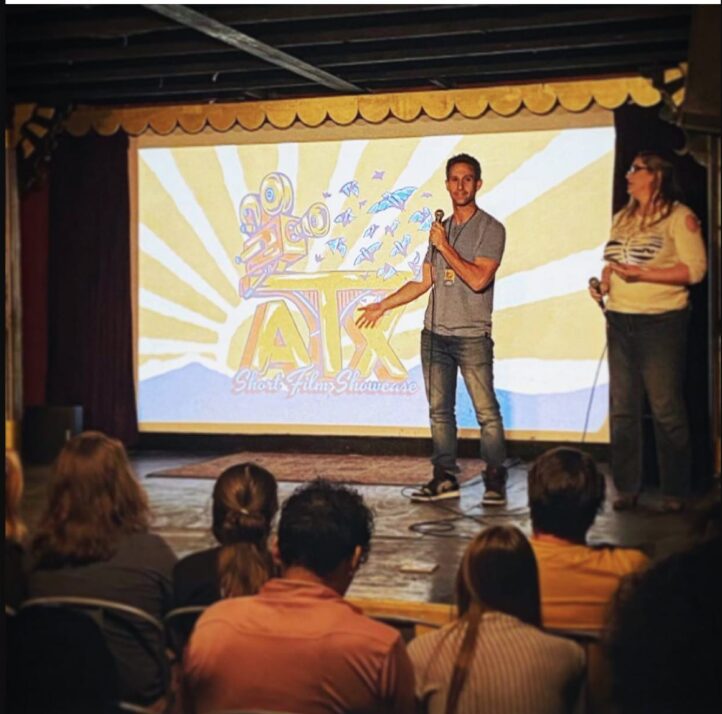 Two people stand on stage in front of a large screen with a colorful logo, audience seated watching.