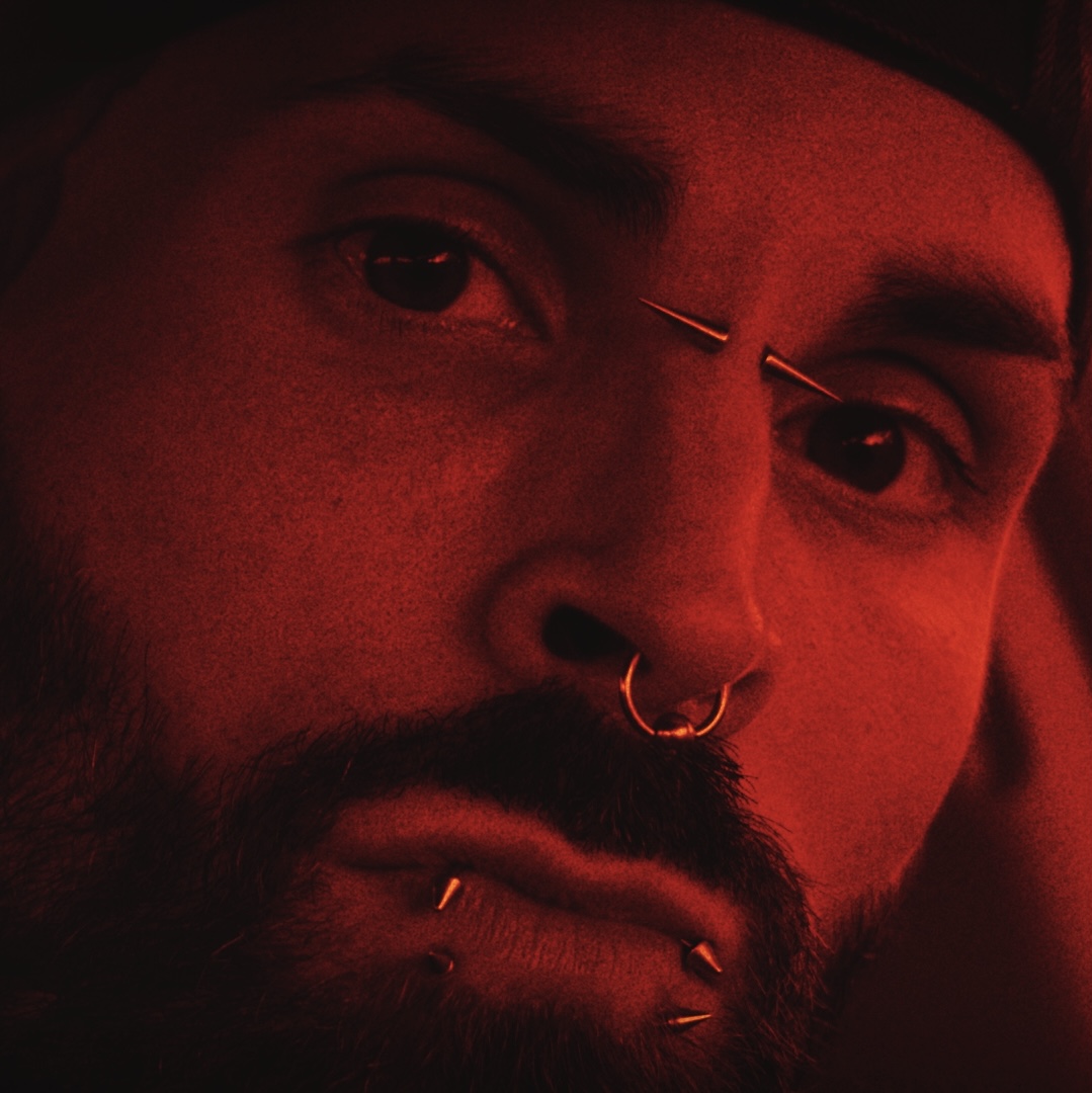Close-up of a person with facial piercings, dark hair, and a beard, illuminated by red lighting.