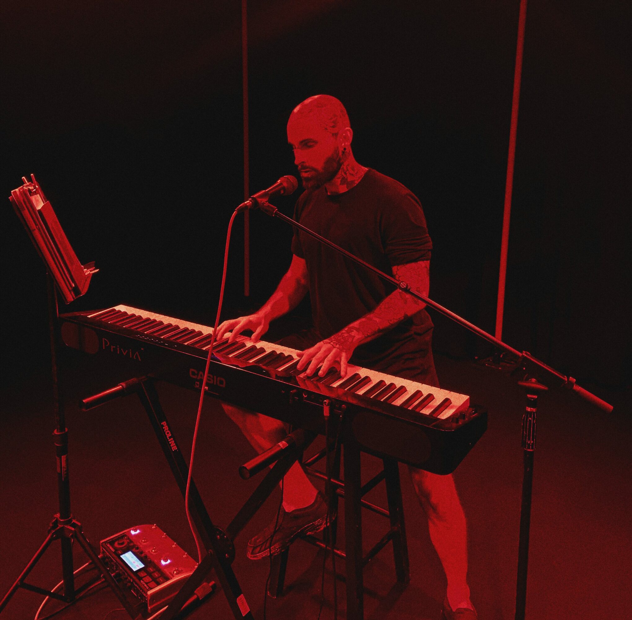 Man playing keyboard on stage with red lighting, microphone, and music stand, standing behind a stool.