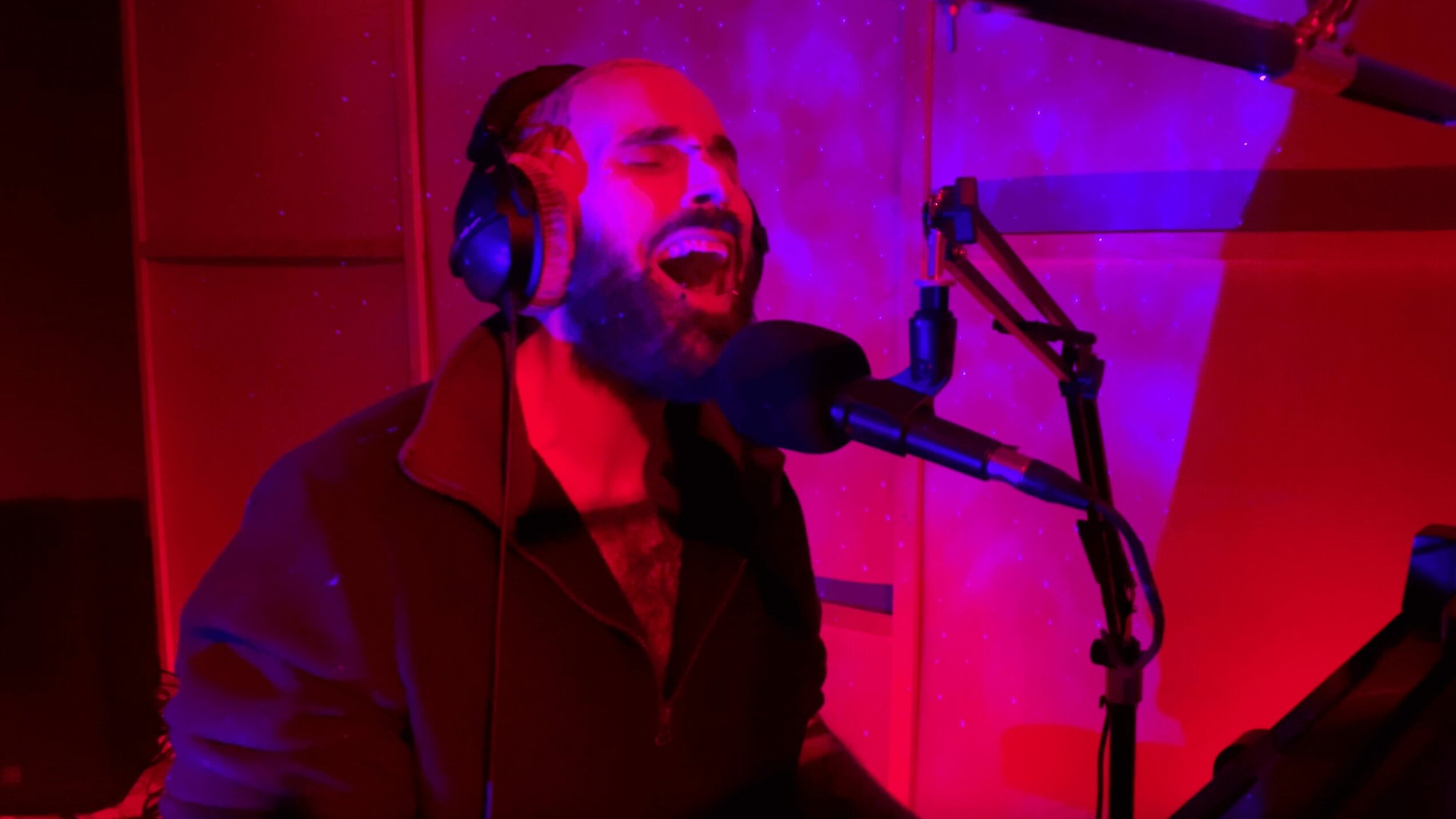 Person singing into a microphone with headphones in a recording studio, illuminated by purple and red lighting.