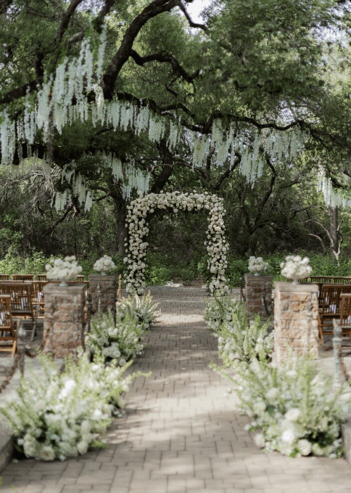 Outdoor wedding setup with an arch decorated with flowers, surrounded by trees and floral arrangements along a brick pathway.