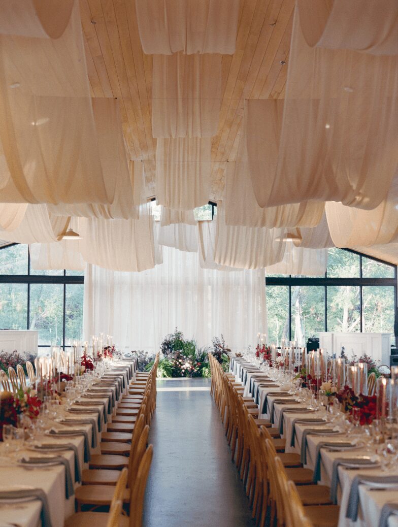 Long banquet tables set with plates, glasses, and floral centerpieces in a bright, airy room with large windows and draped ceiling.