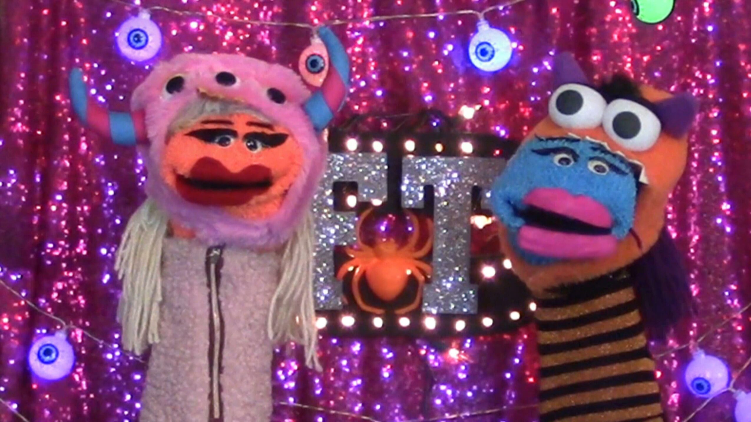 Two puppets with colorful costumes and large eyes stand in front of a sparkly purple background with lights and a crab decoration.