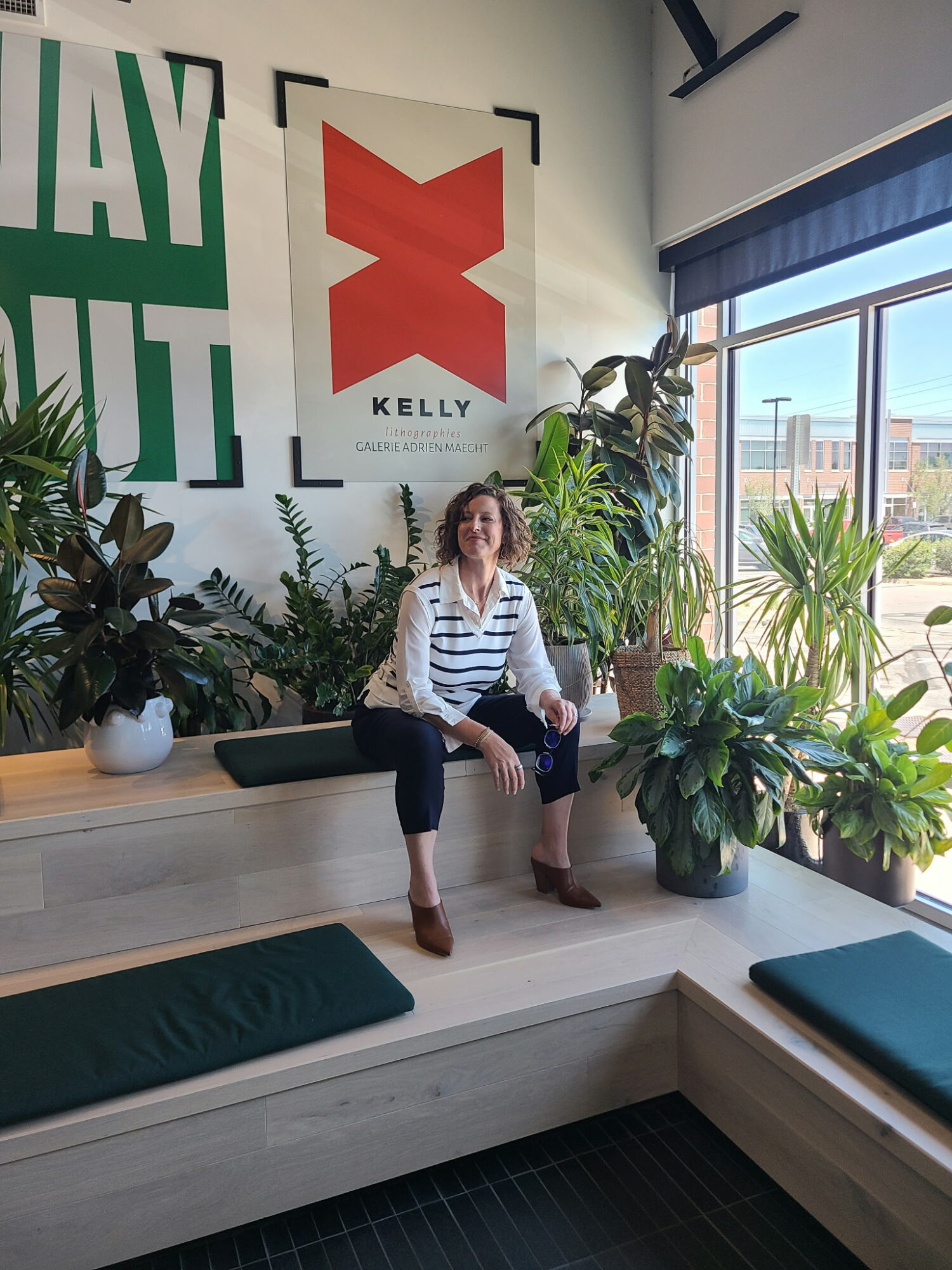 Woman sitting on a wooden bench surrounded by plants, with large windows and posters on the wall behind her.