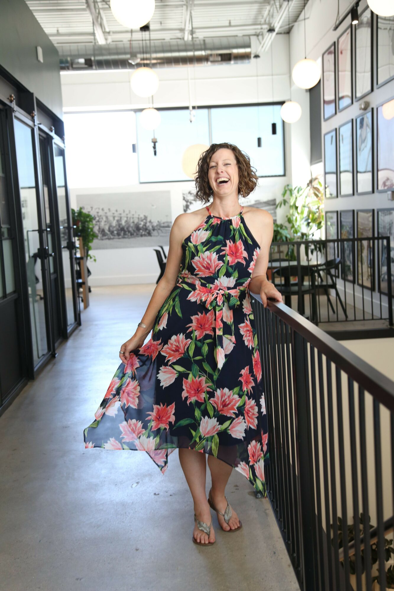 Woman in floral dress standing on a balcony with railing, indoor space with large windows and hanging lights behind her.