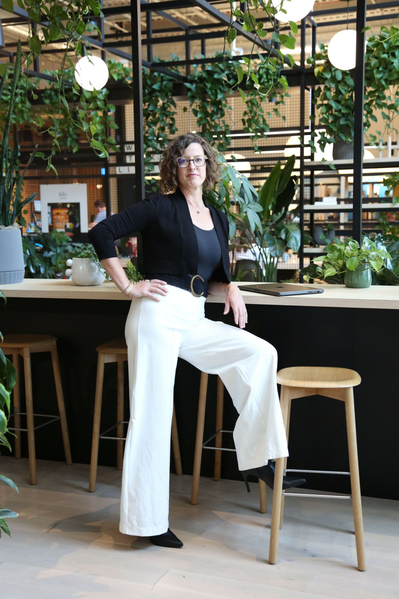 Person with curly hair in black top and white pants standing indoors near plants and wooden furniture.