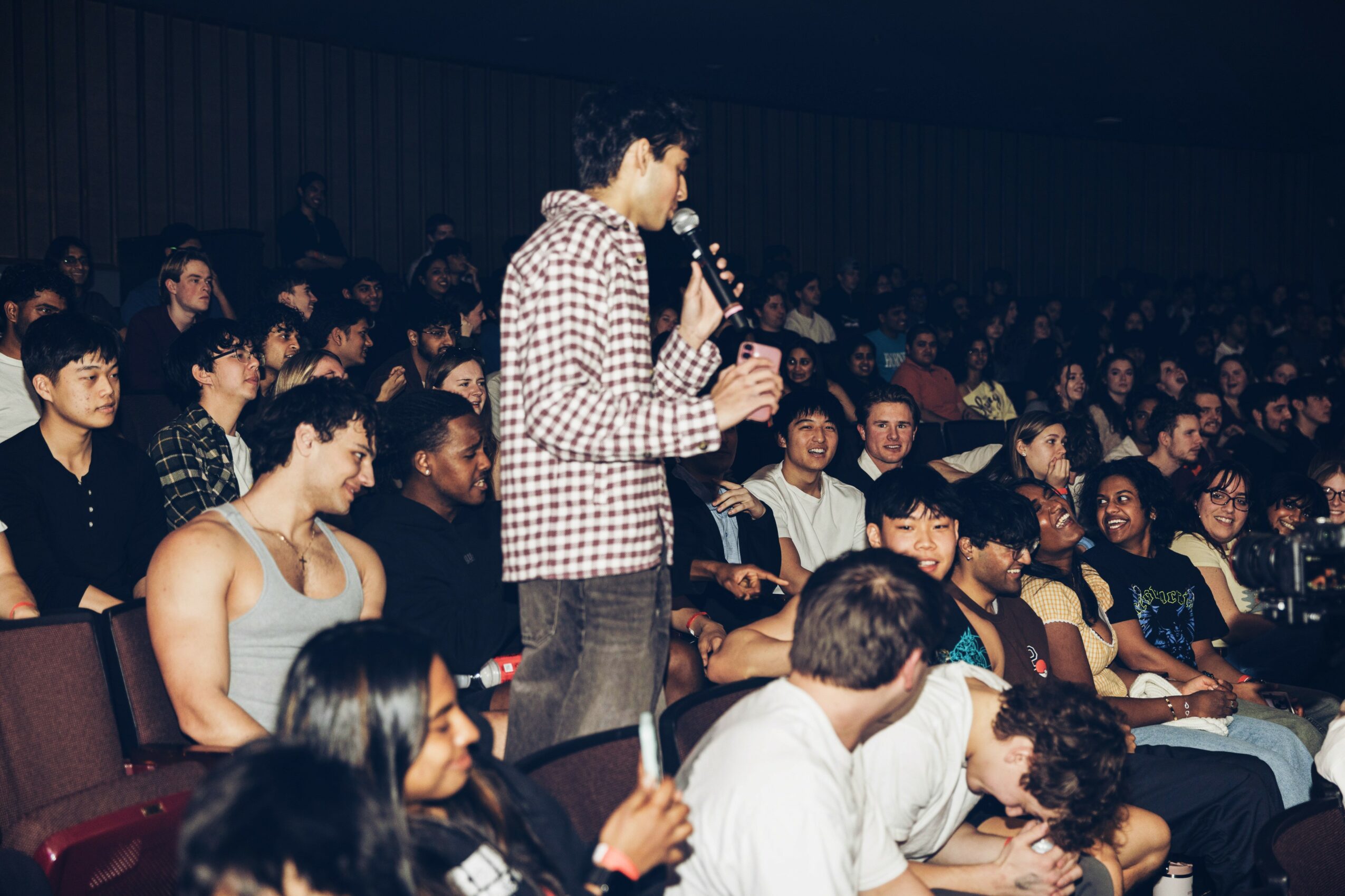 Person standing and speaking into a microphone in front of a seated audience in a large indoor space.