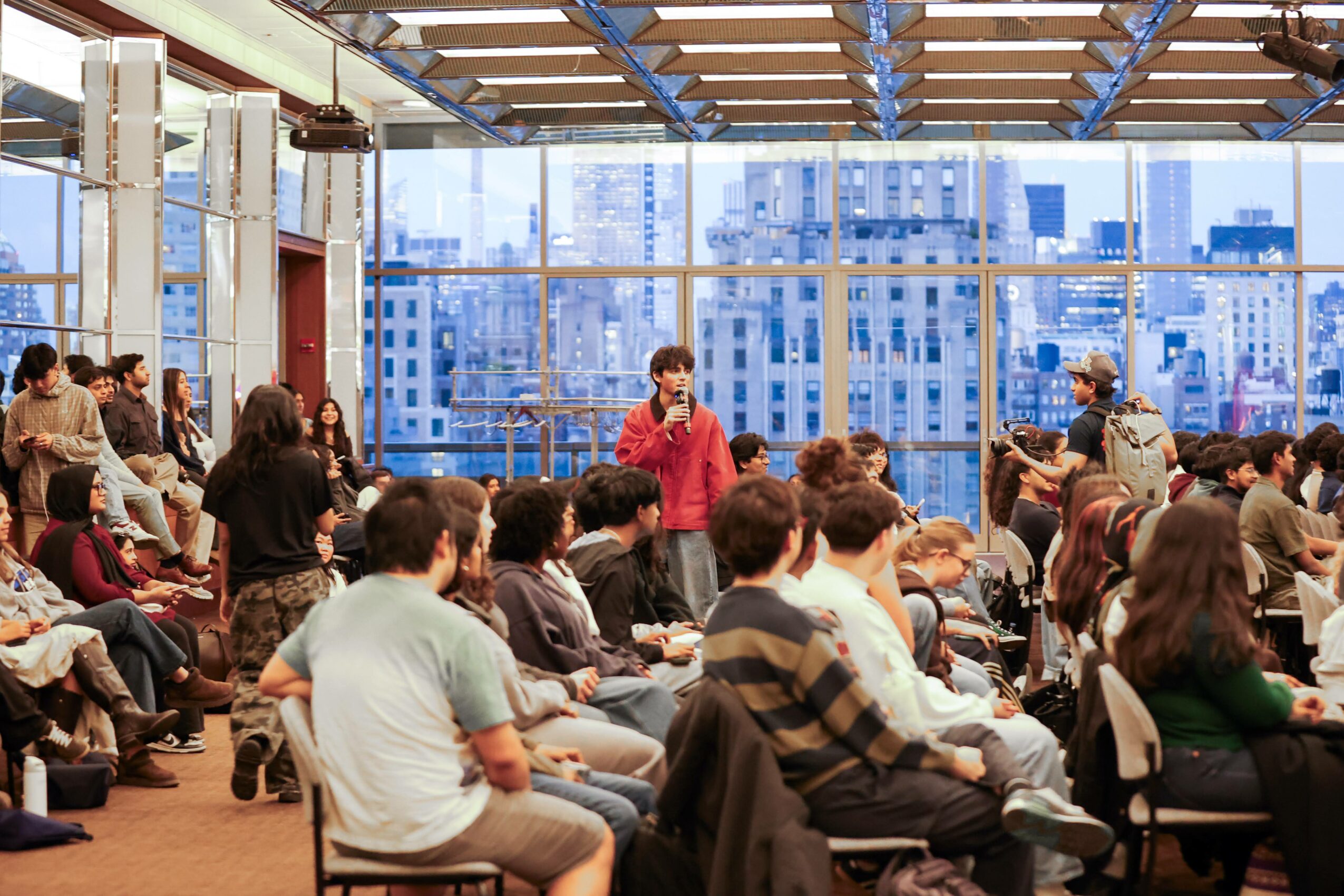 People seated in a large room with floor-to-ceiling windows showing a cityscape, some standing and talking, others listening.