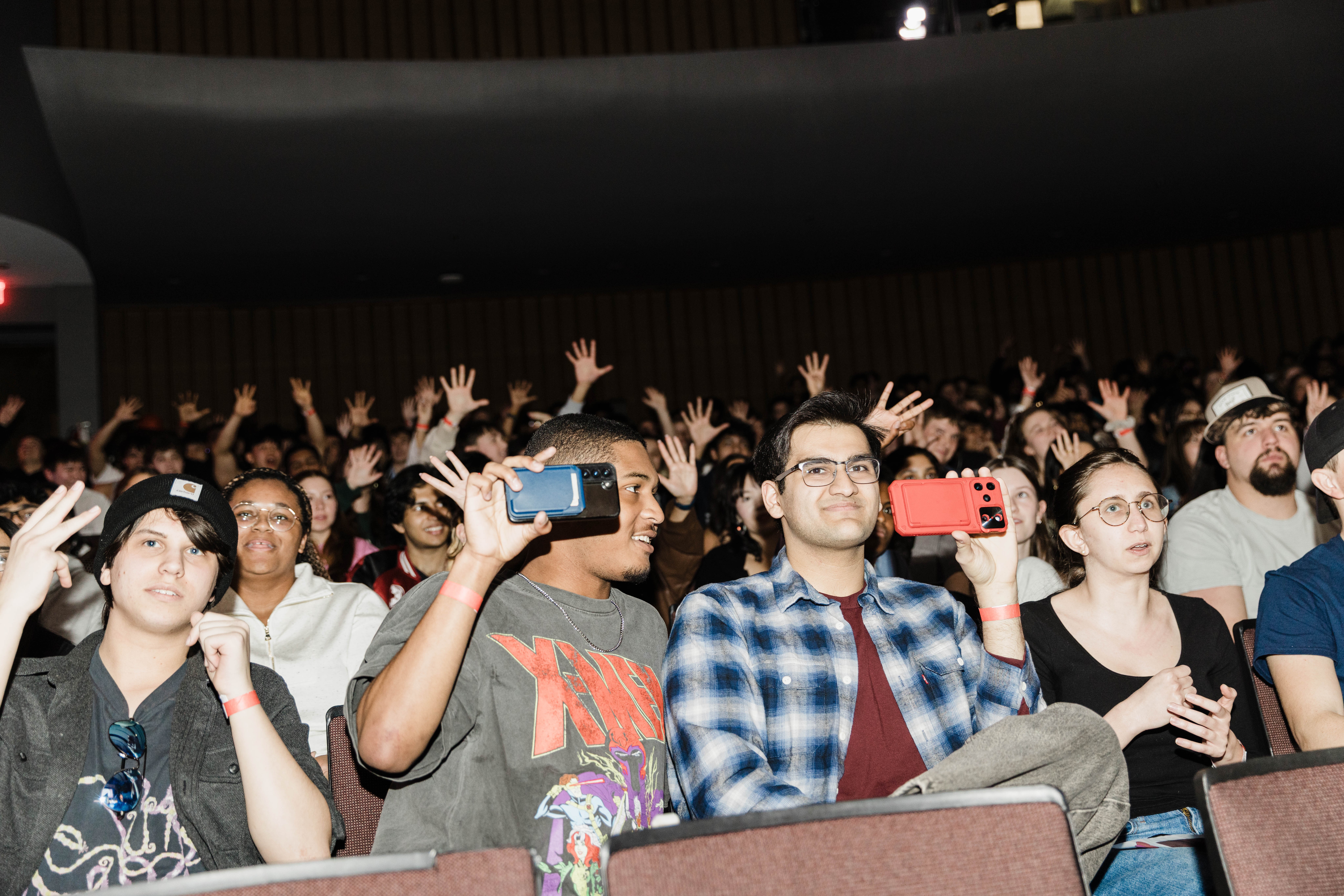 Crowd of people sitting and standing in an indoor event, some taking photos or videos with smartphones, diverse ages and styles.