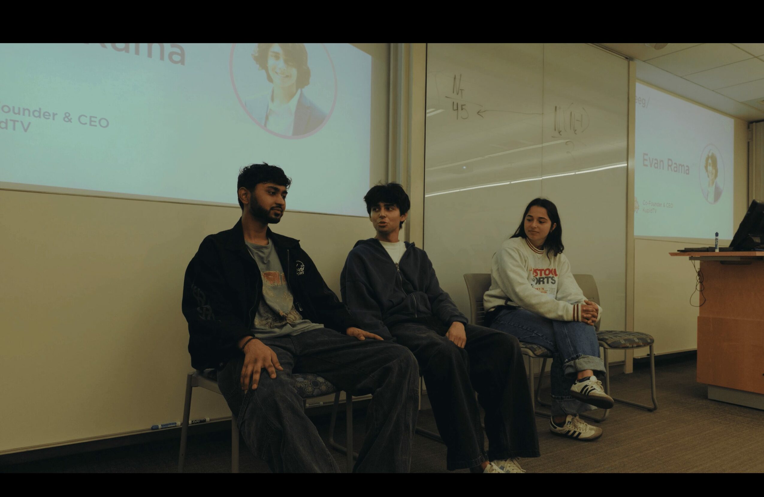 Three people sitting on chairs in a classroom, with a presentation screen behind them, and a whiteboard on the right.
