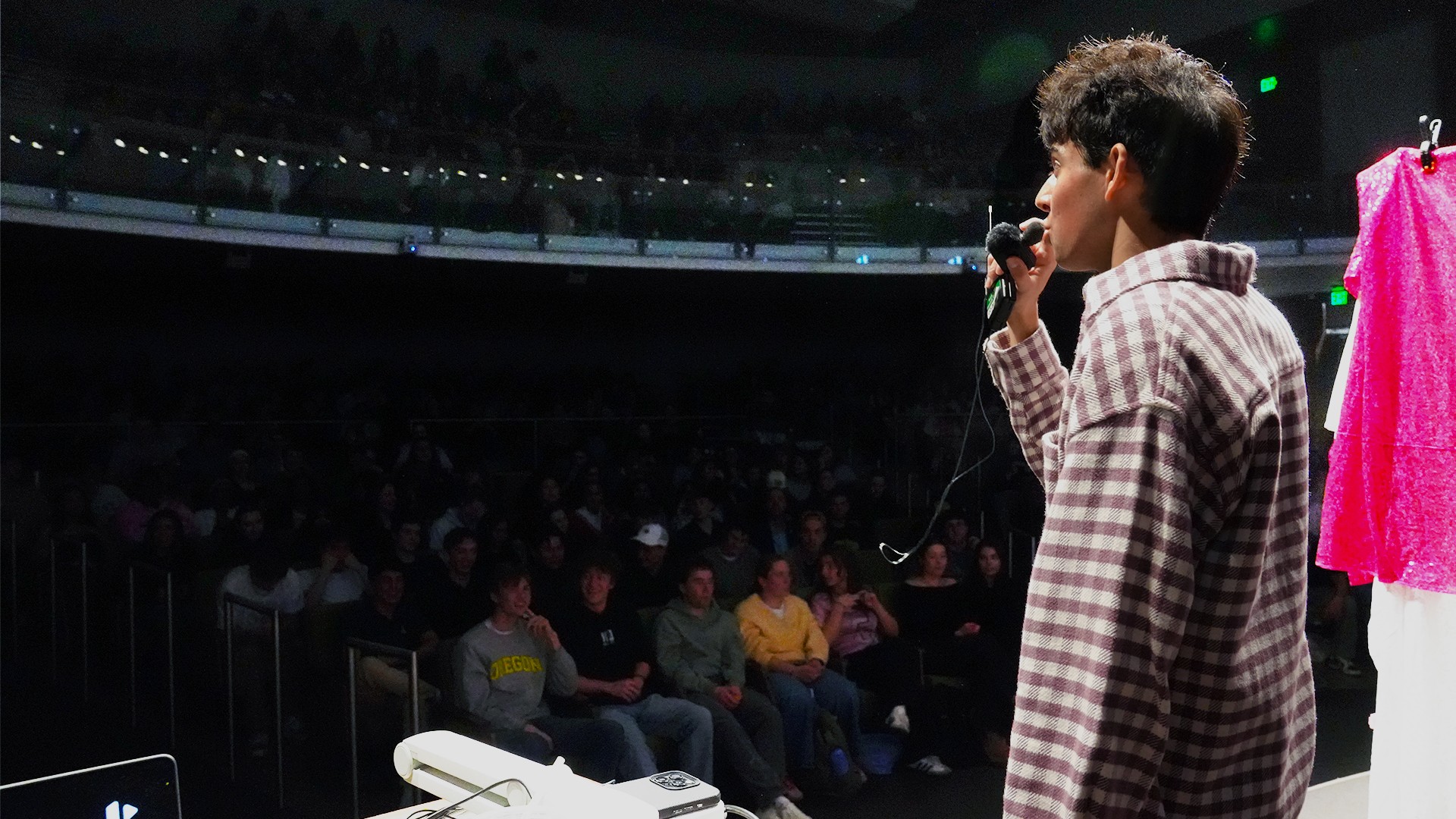 Person speaking into microphone on stage with audience in background, pink cloth hanging nearby.