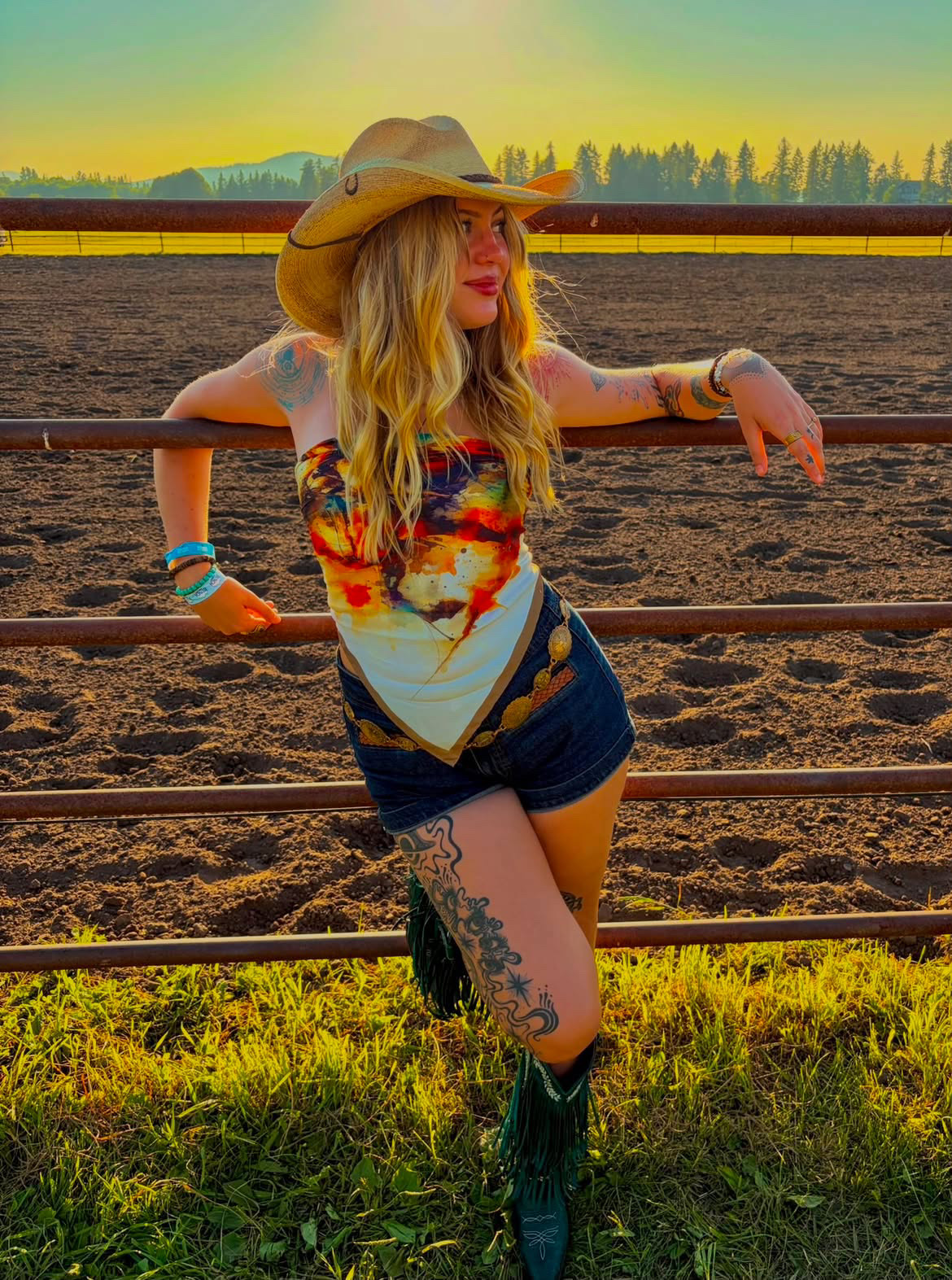 Young woman with long blonde hair wearing a cowboy hat, colorful top, and shorts, leaning on a fence outdoors.