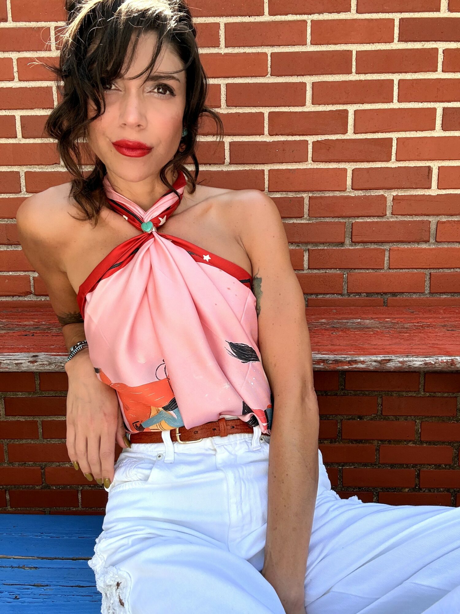 Woman with dark wavy hair and red lipstick, wearing a pink and red top, sitting against a brick wall.
