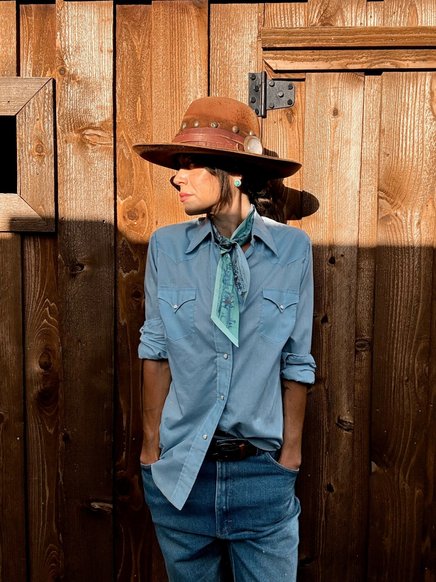 Woman wearing a wide-brimmed hat, blue shirt, and jeans stands against a wooden fence, looking to the side.