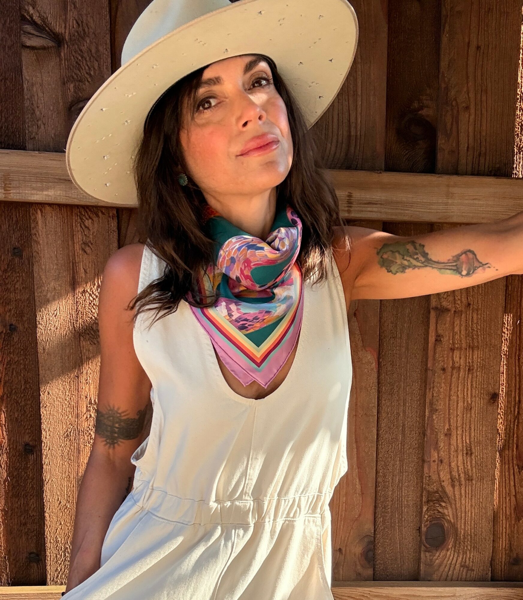 Woman wearing a wide-brimmed hat and colorful scarf, standing against a wooden fence, with tattoos on her arms.