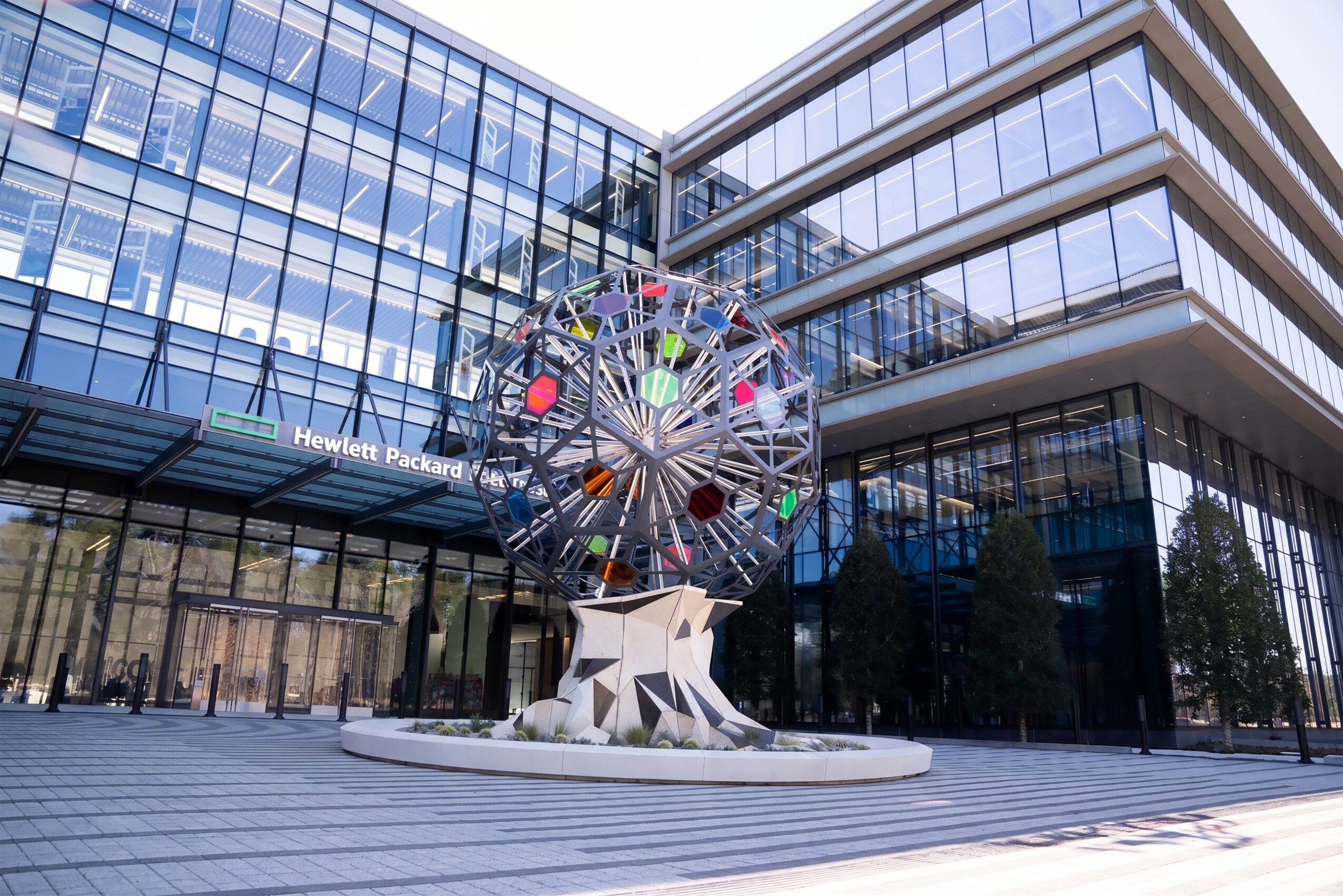 Modern glass building with a colorful Ferris wheel sculpture in front, surrounded by paved area and trees.