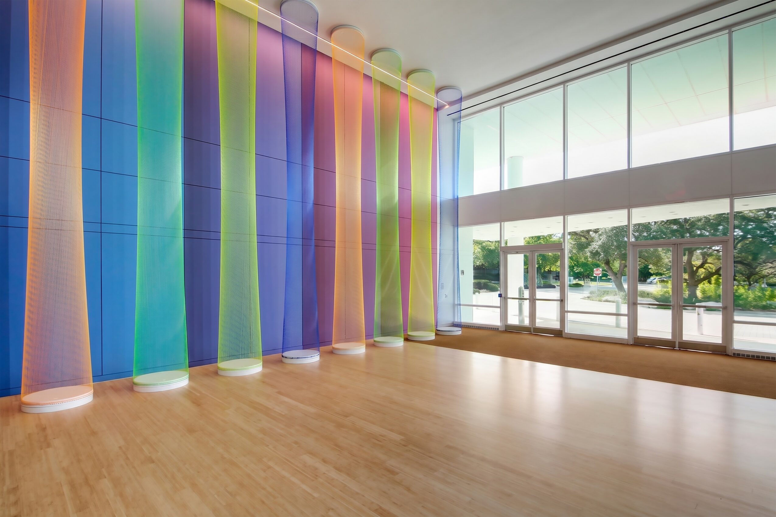 Interior space with colorful vertical curtains, large windows, and wooden floor, bright natural light, and outdoor greenery visible.