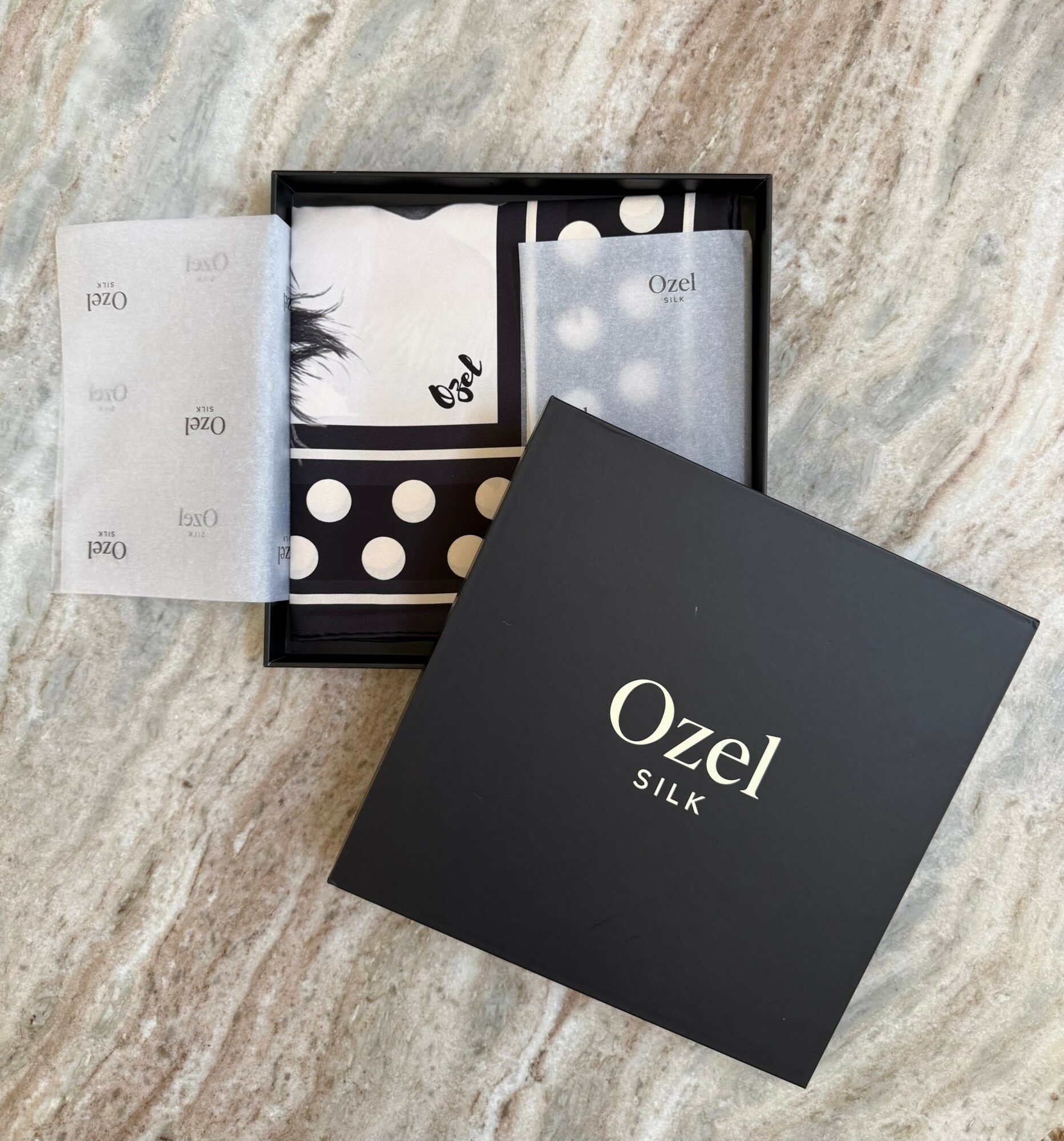 Open black box with silk accessories and packaging on a marble surface, with a black box lid nearby.