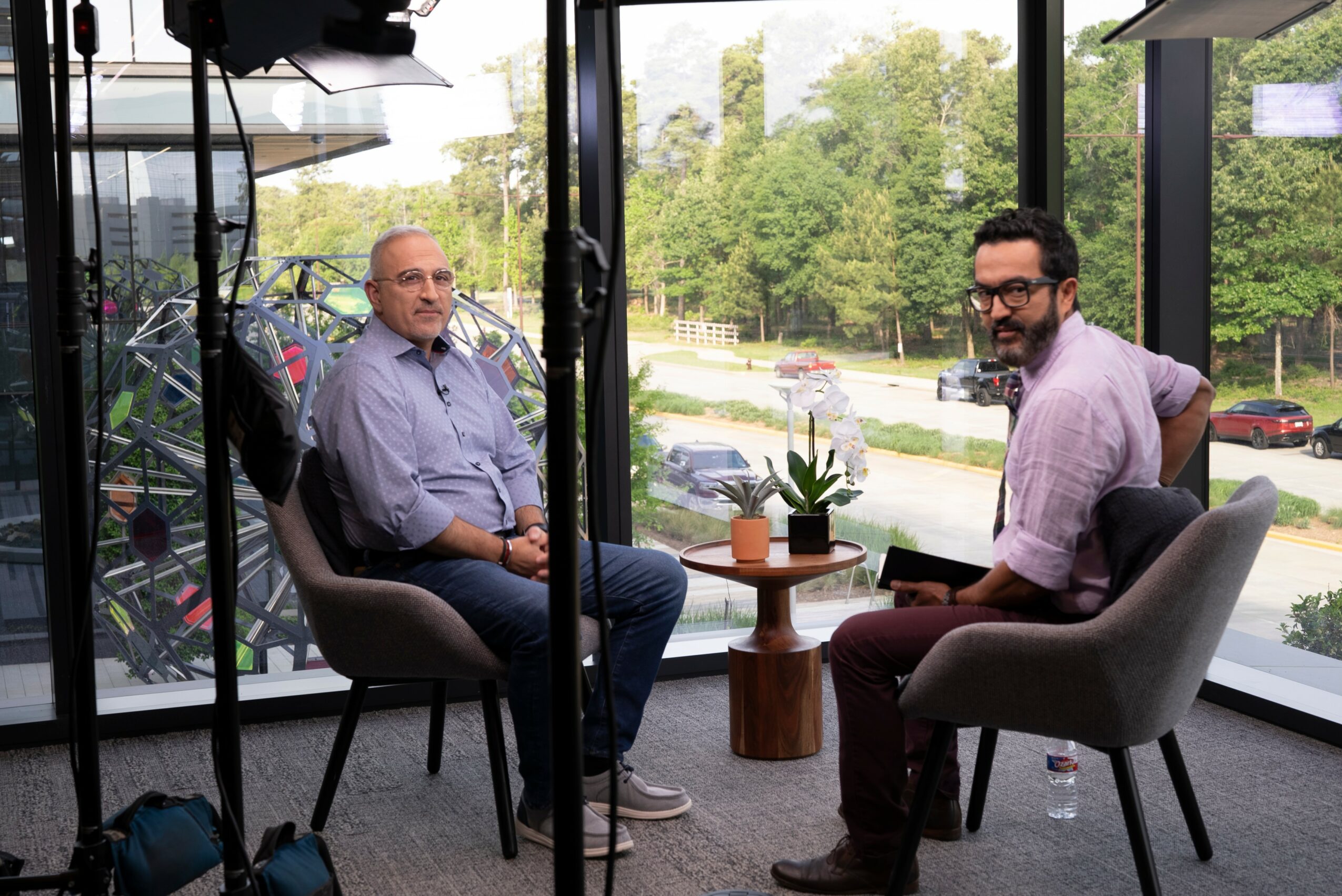 Two men sit in chairs inside a room with large windows, greenery and cars visible outside, during daytime.