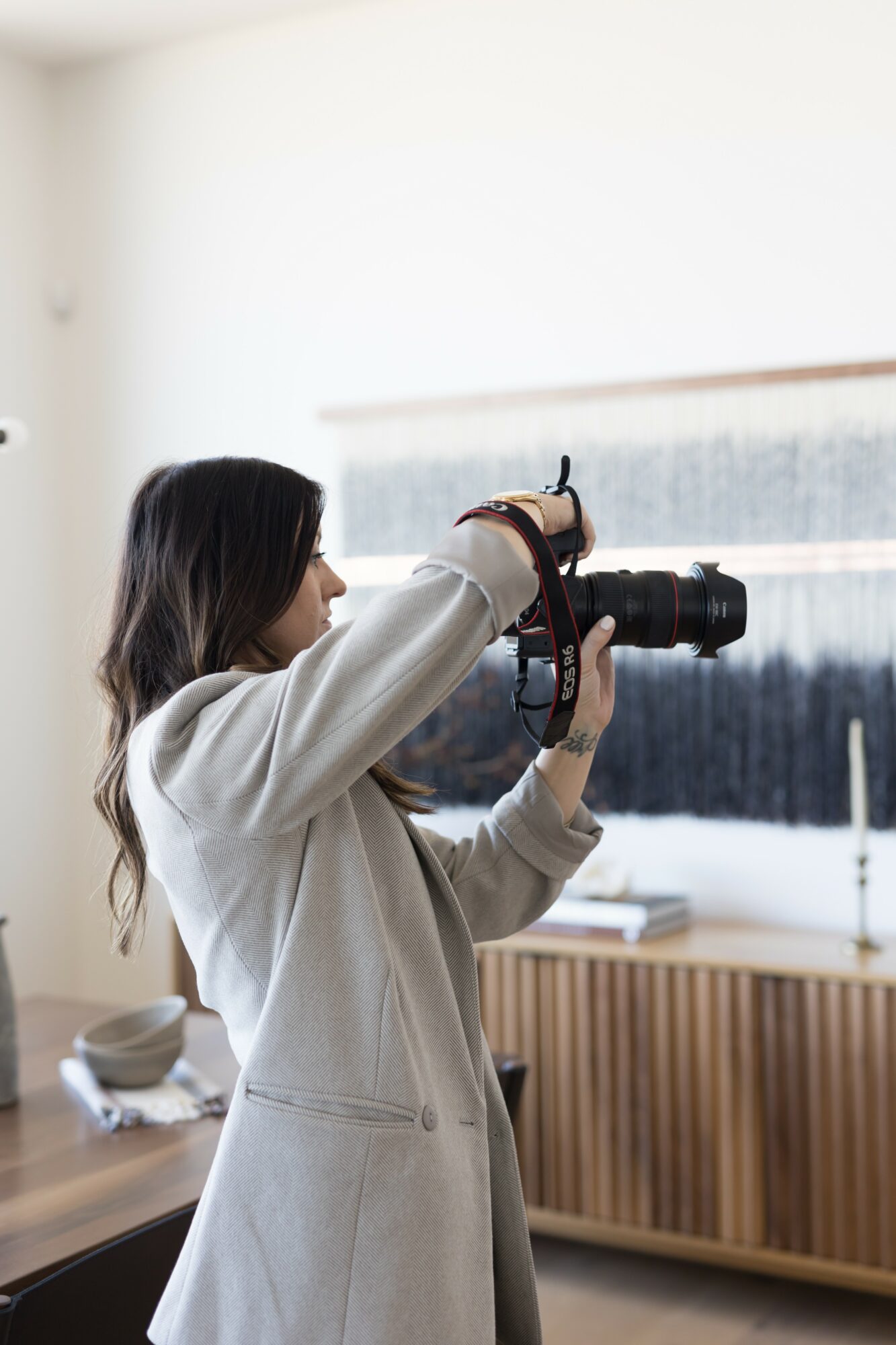 Person with long hair holding a camera, looking out a window with a snowy landscape, wearing a light-colored coat.