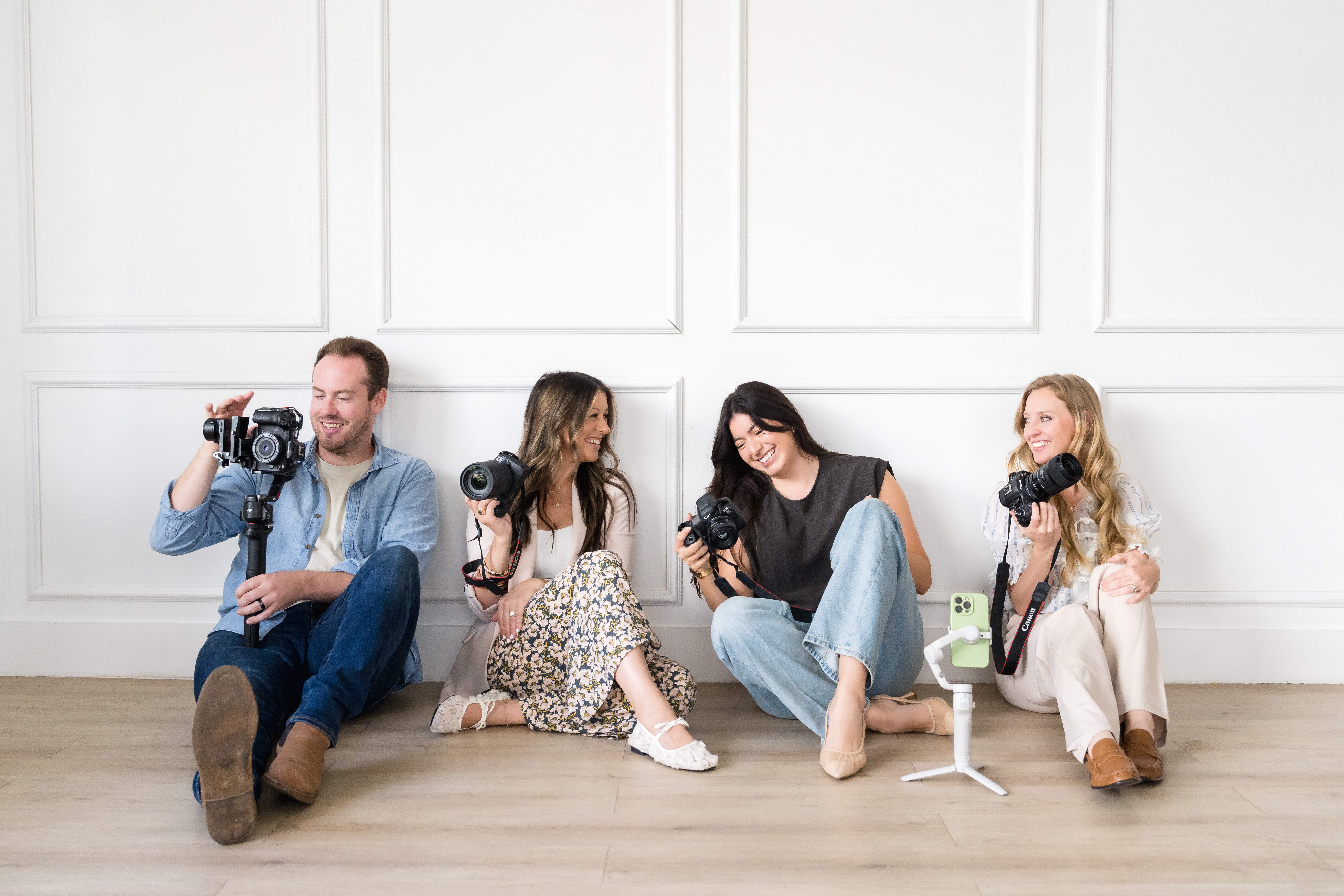 Four people sitting on the floor against a white wall, holding cameras and smiling, with a wooden floor.