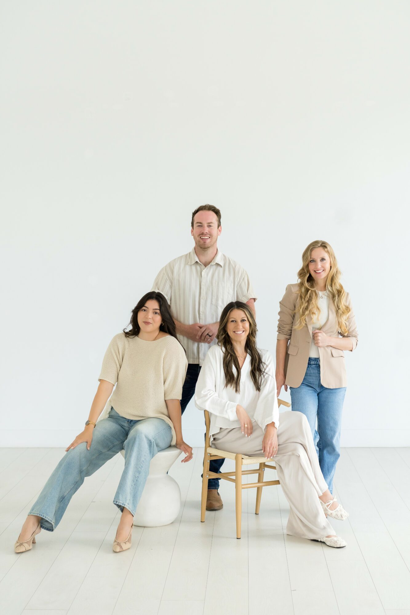 Four people pose against a plain white background, with one seated on a chair and others standing or sitting nearby.