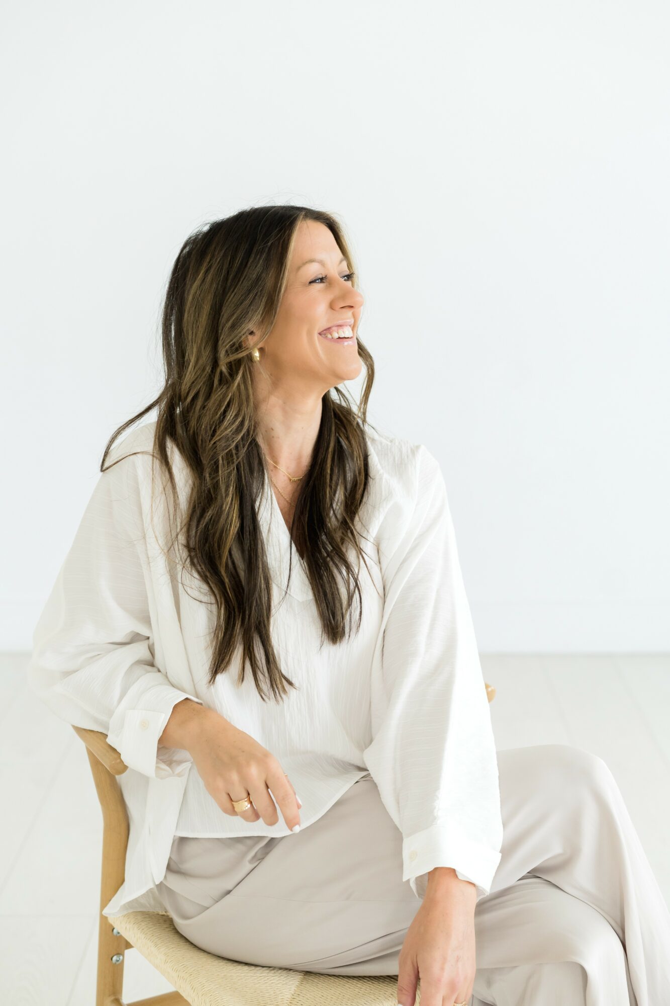 Woman with long wavy hair smiling, sitting on a chair against a plain white background.