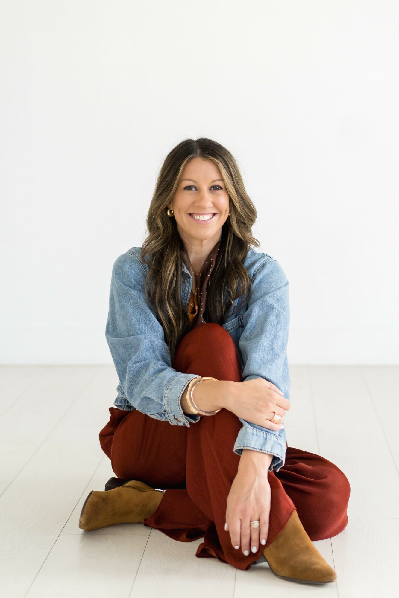 Woman sitting on the floor smiling, wearing a denim jacket, maroon pants, and brown boots, against a white background.