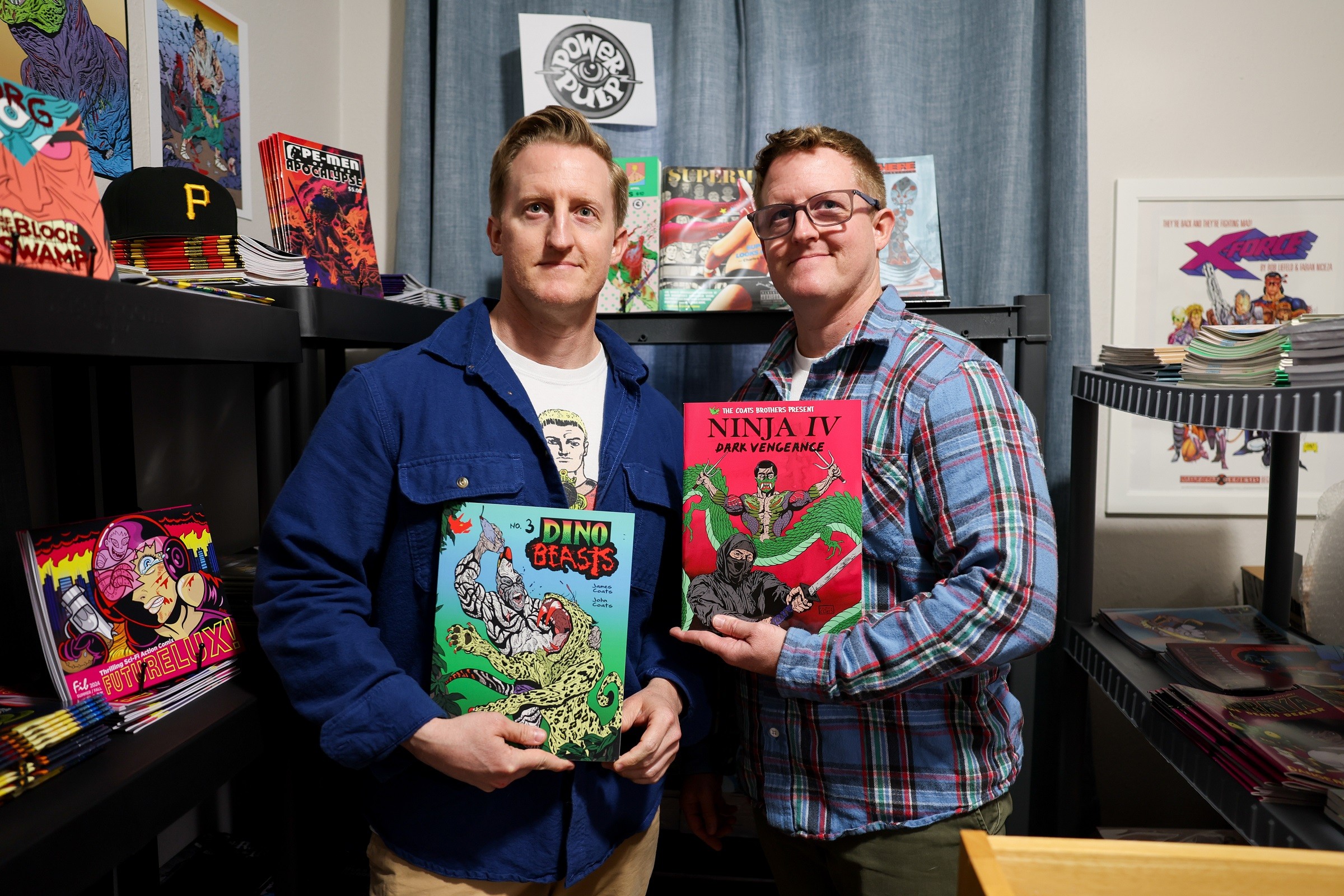 Two men holding colorful comic books in a room with shelves of books and artwork, smiling at the camera.