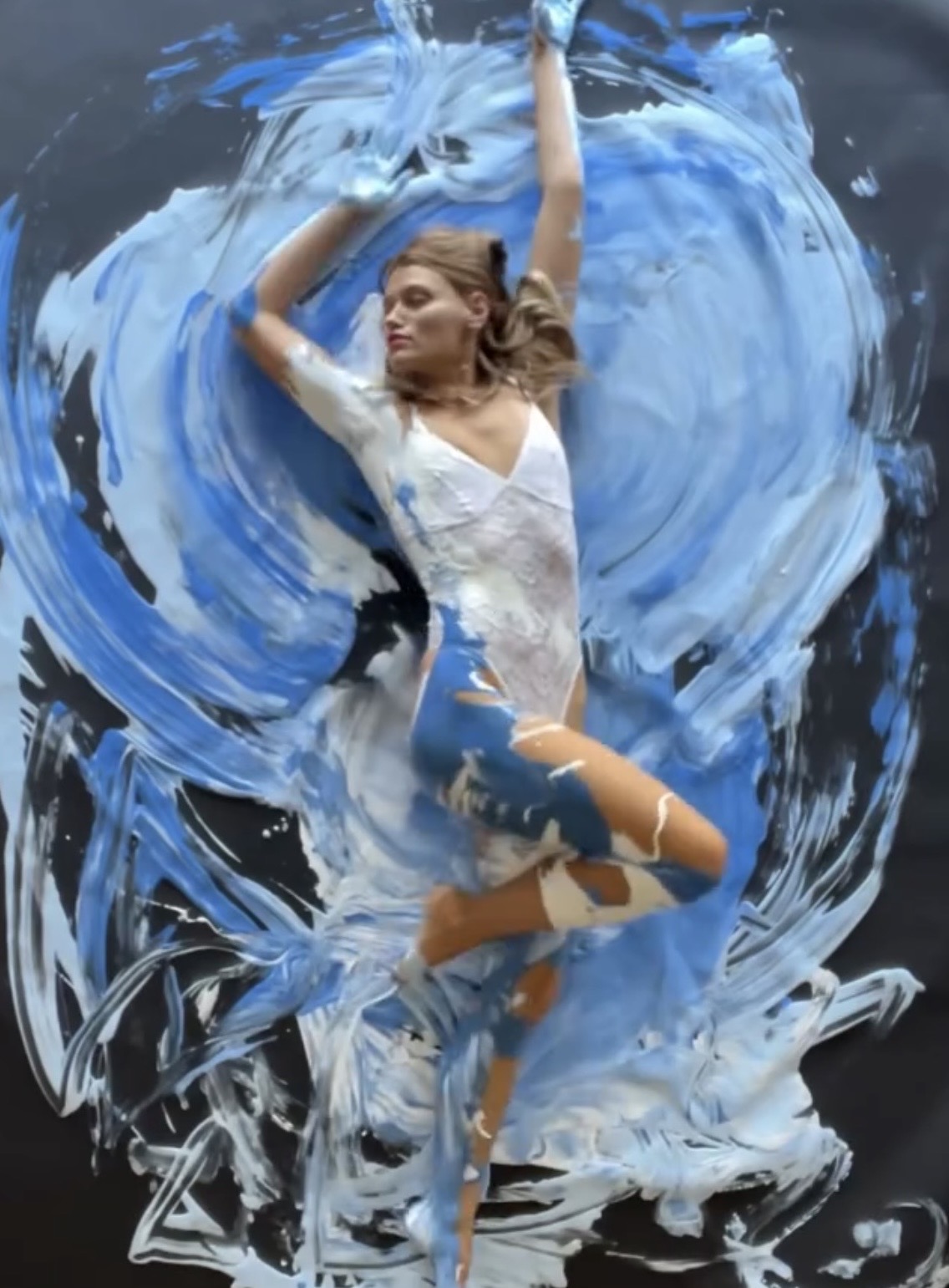 Woman floating in water with blue and white paint splashes around her, arms raised above her head, wearing a white top and shorts.