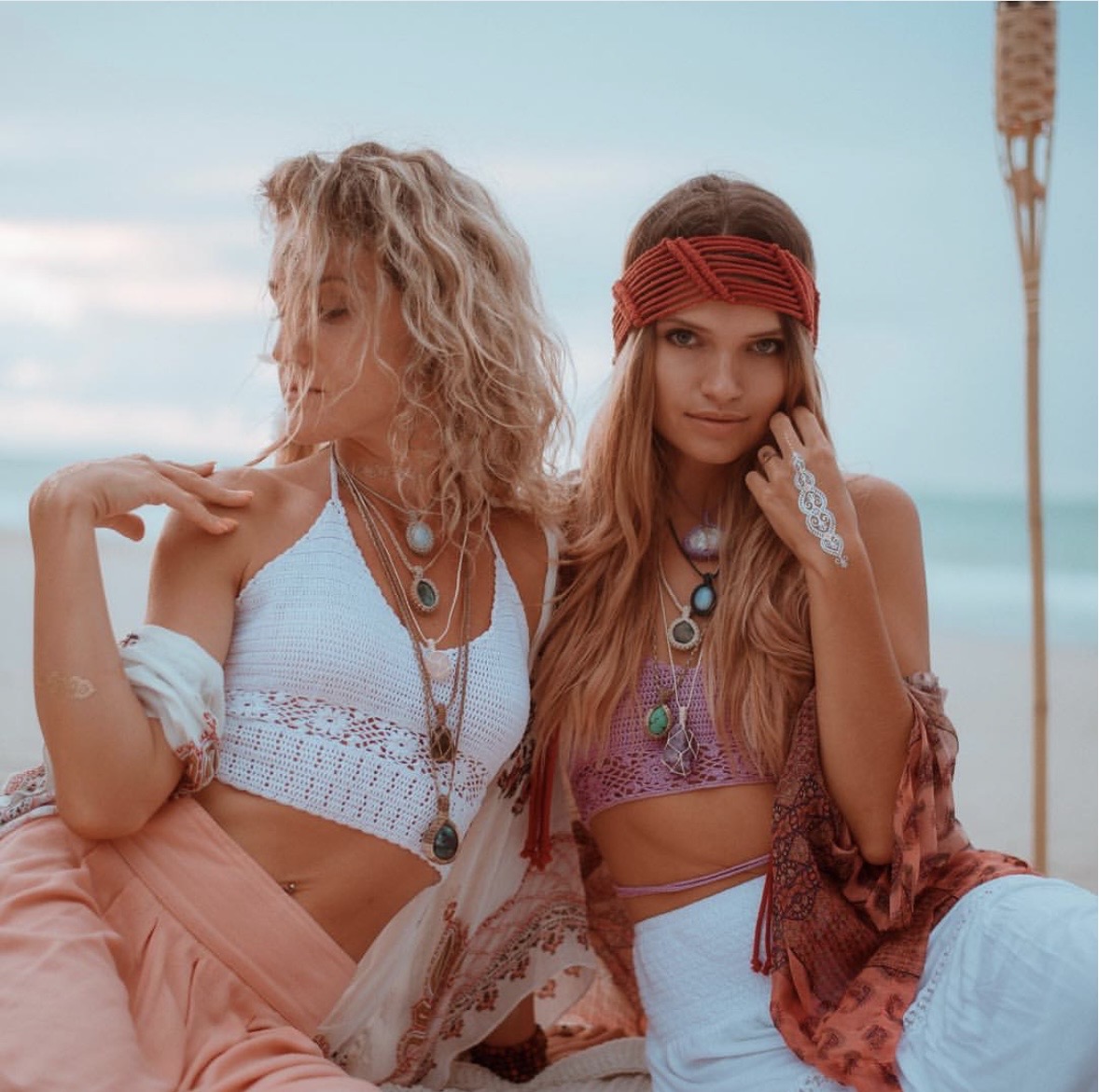 Two women sit on a beach, one with curly blonde hair and the other with long straight hair, wearing bohemian clothing and accessories.