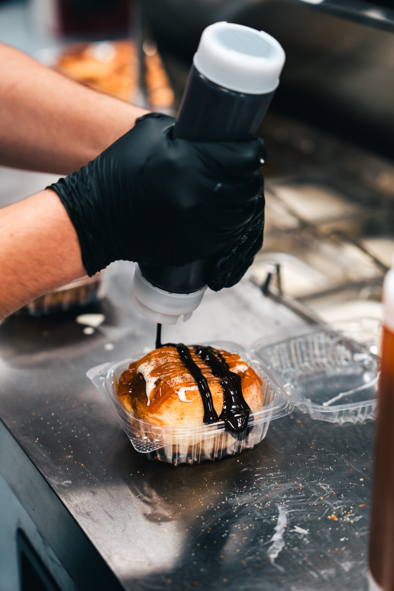 Person wearing black gloves applies sauce to a pastry with a squeeze bottle, pastry wrapped in plastic on a metal surface.