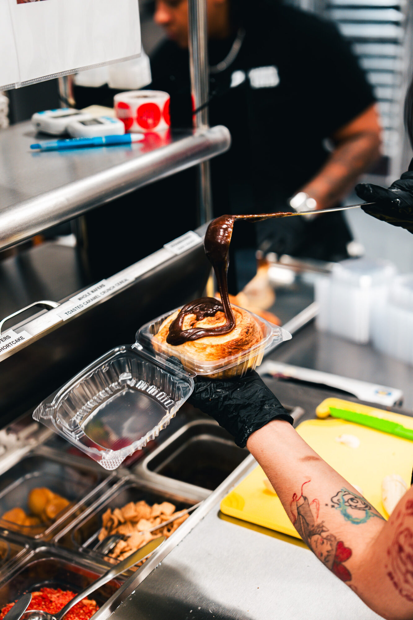 Person pouring chocolate over a pastry in a kitchen, with a tattooed arm and various kitchen tools visible.