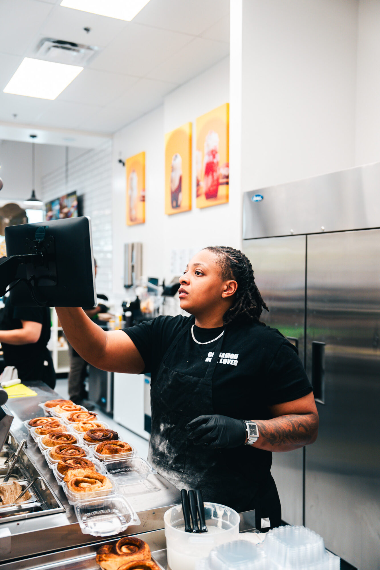 A woman in a black uniform prepares food in a commercial kitchen, with pastries on a tray in front of her.